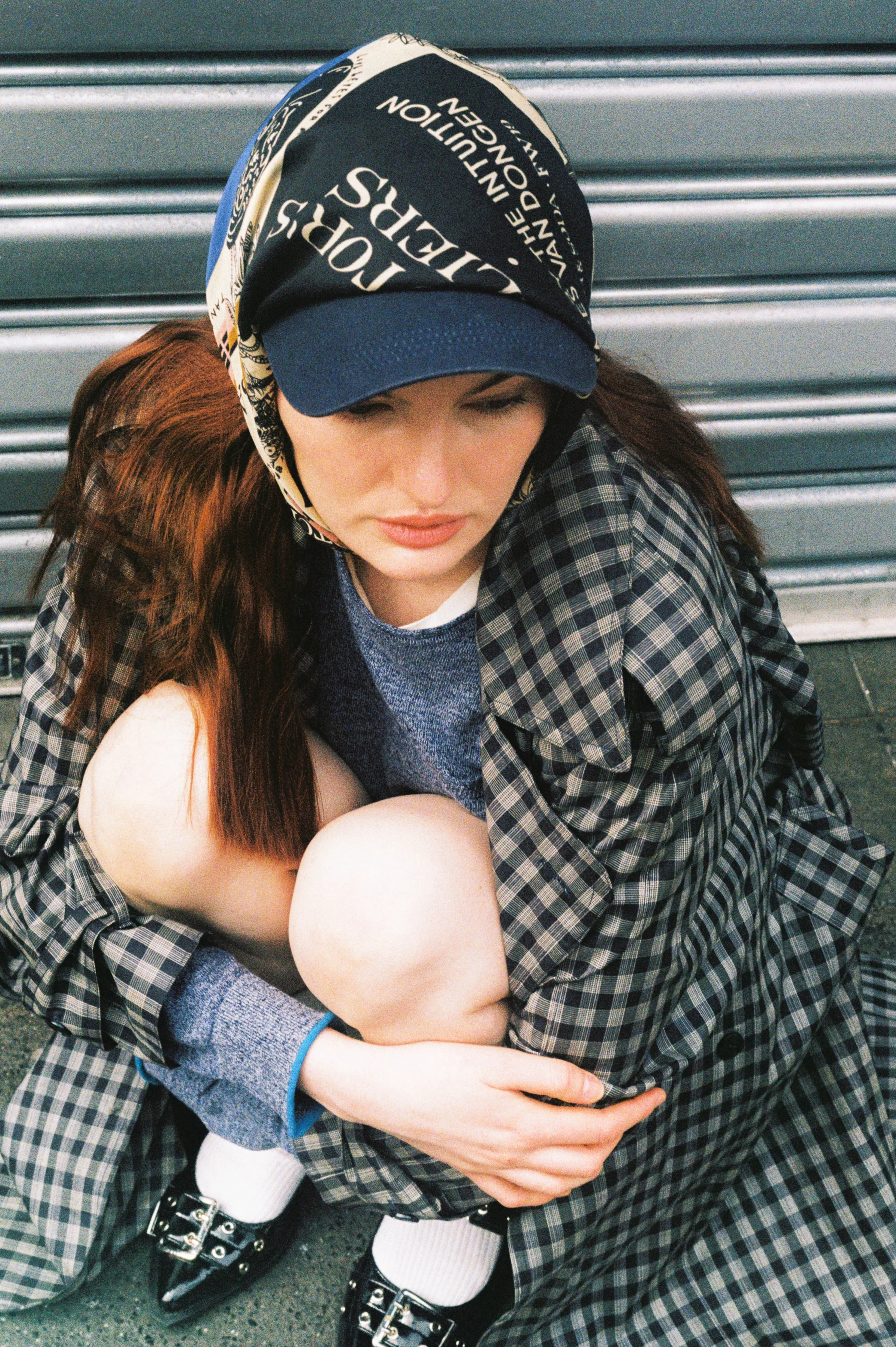 A young woman with long red hair, wearing a newsboy cap, plaid coat, and socks with black shoes, is sitting on the ground with her knees up and eyes closed, in front of a metal shutter.