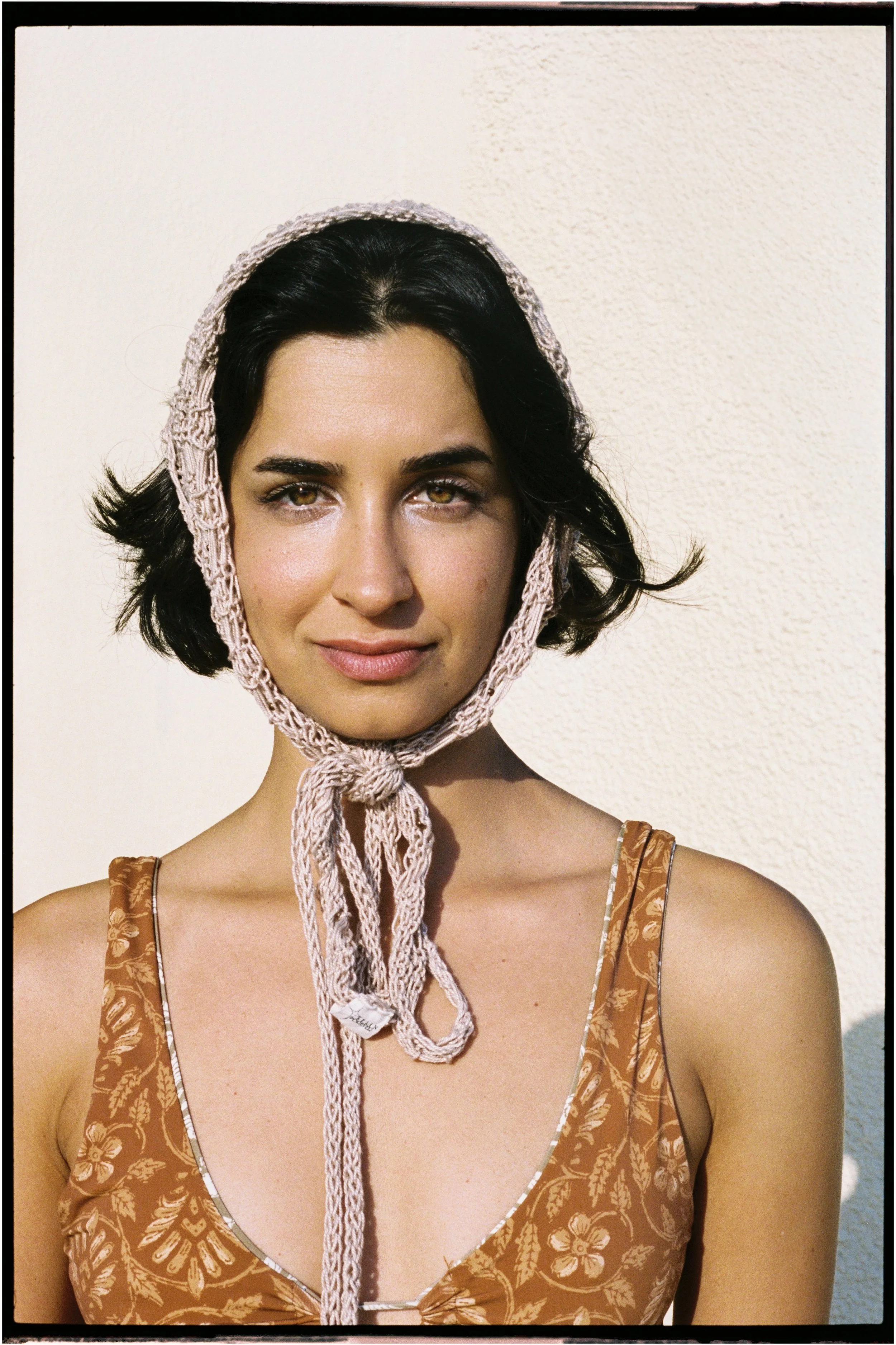 Portrait of a young woman with dark hair, wearing a crocheted hood tied under her chin, and a floral-patterned sleeveless dress, standing against a textured light background.