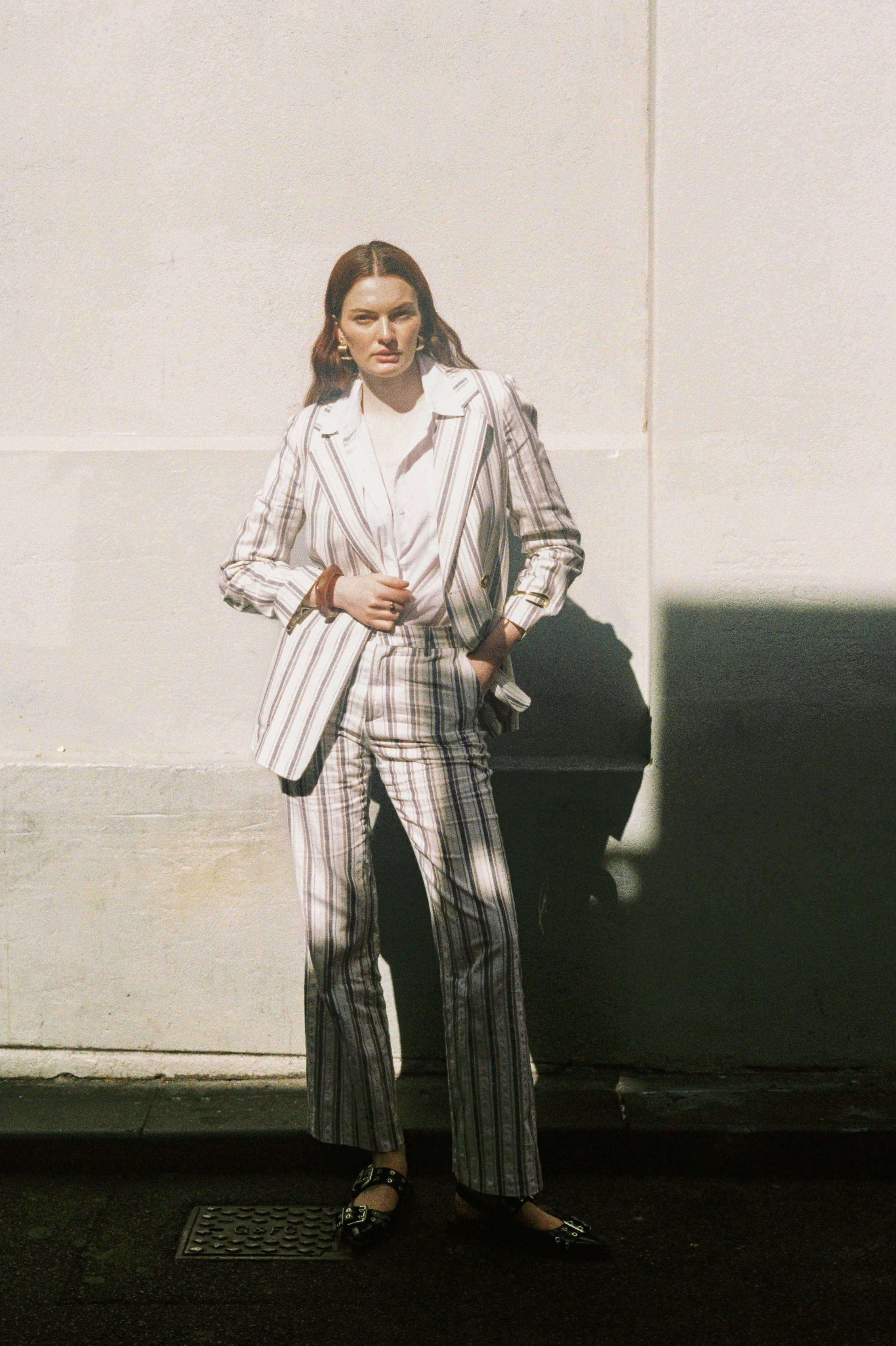 A woman in a striped suit standing against a white wall, casting a shadow, with her hand on her waist.