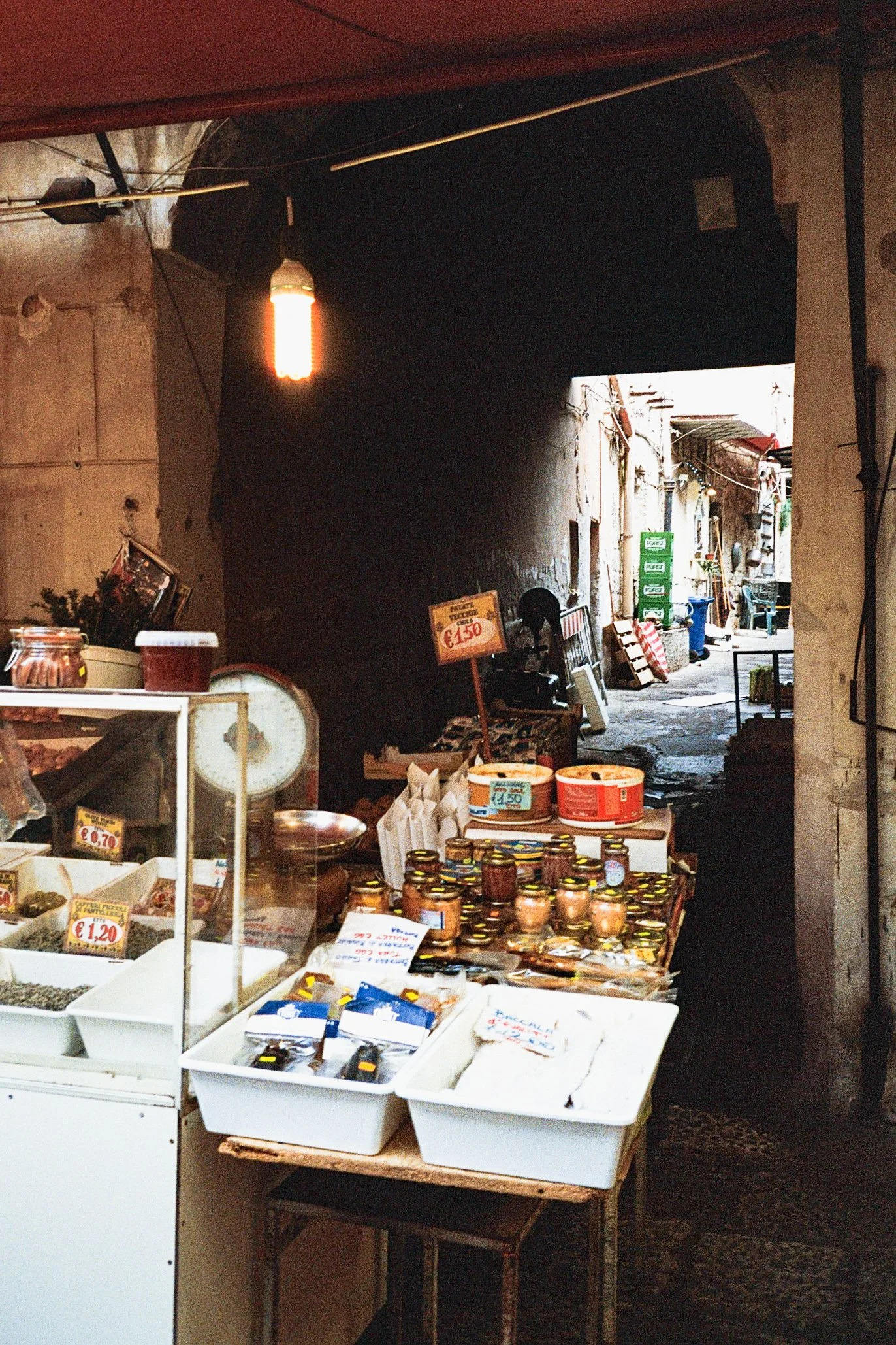 A small marketplace stall displaying jars of jams and containers of spices or herbs in a narrow alleyway.