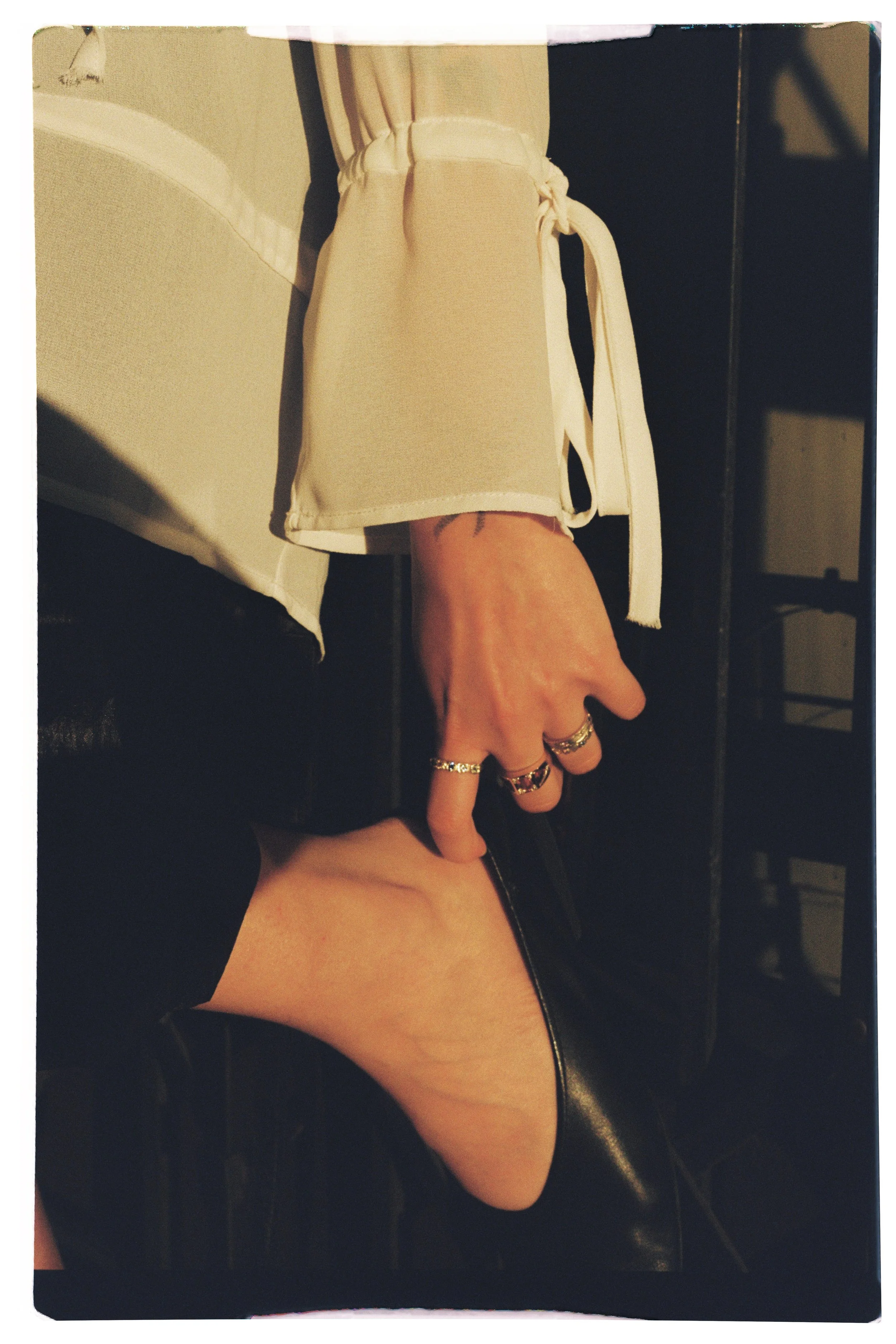 Person sitting with her foot resting on a black leather chair, wearing black shoes, beige pants, a beige shirt, and multiple rings on her fingers.
