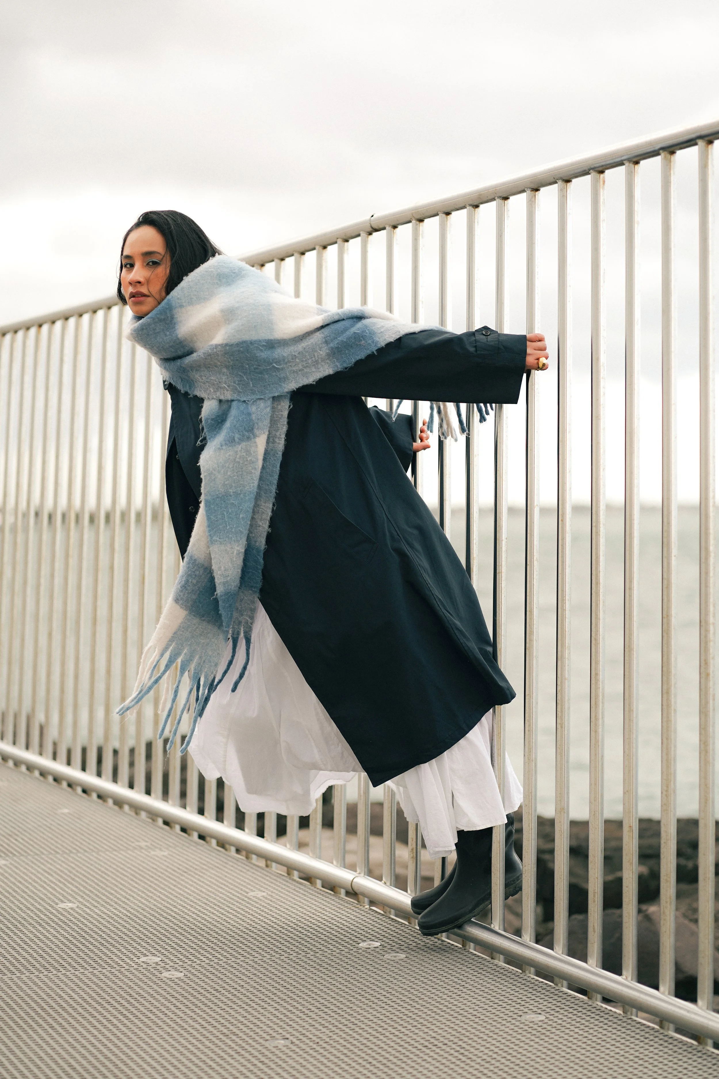 Woman leaning on a metal railing outdoors, wearing a black coat, white skirt, black boots, and a large blue and white checkered scarf.