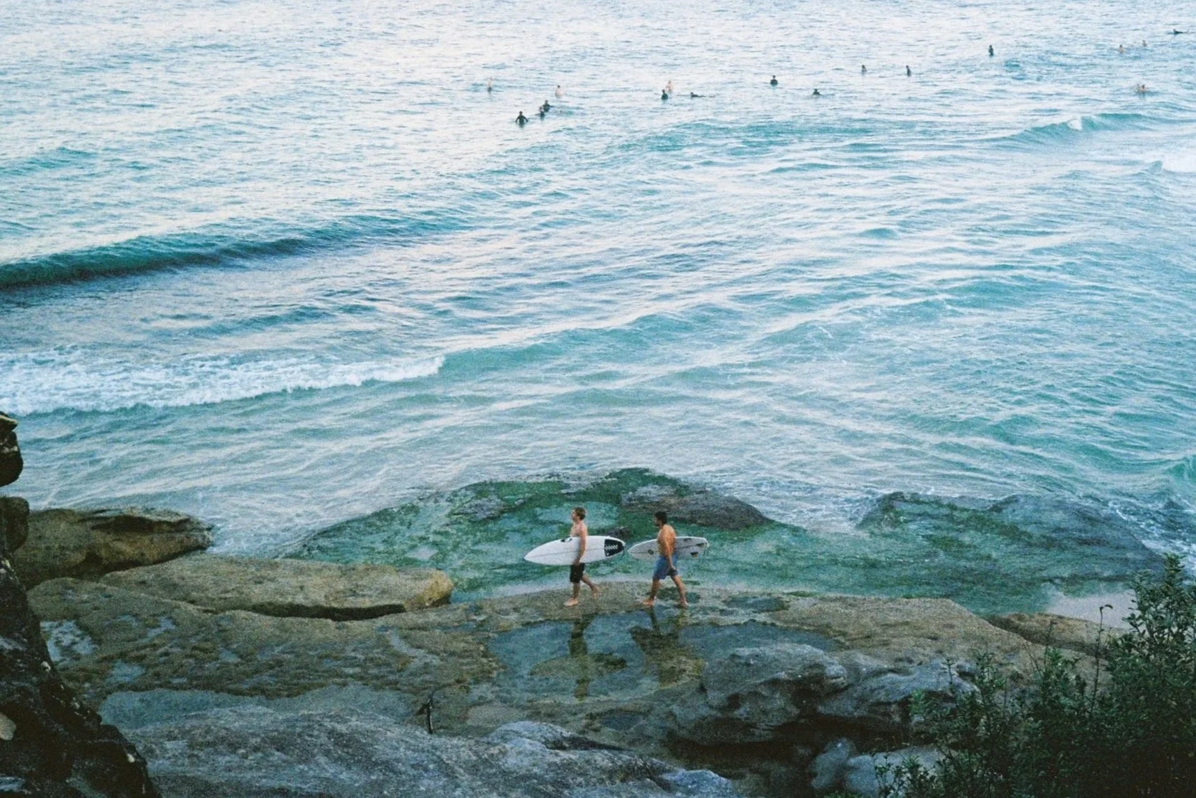 Two surfers walking along a rocky shoreline carrying surfboards, with the ocean and other surfers in the background.