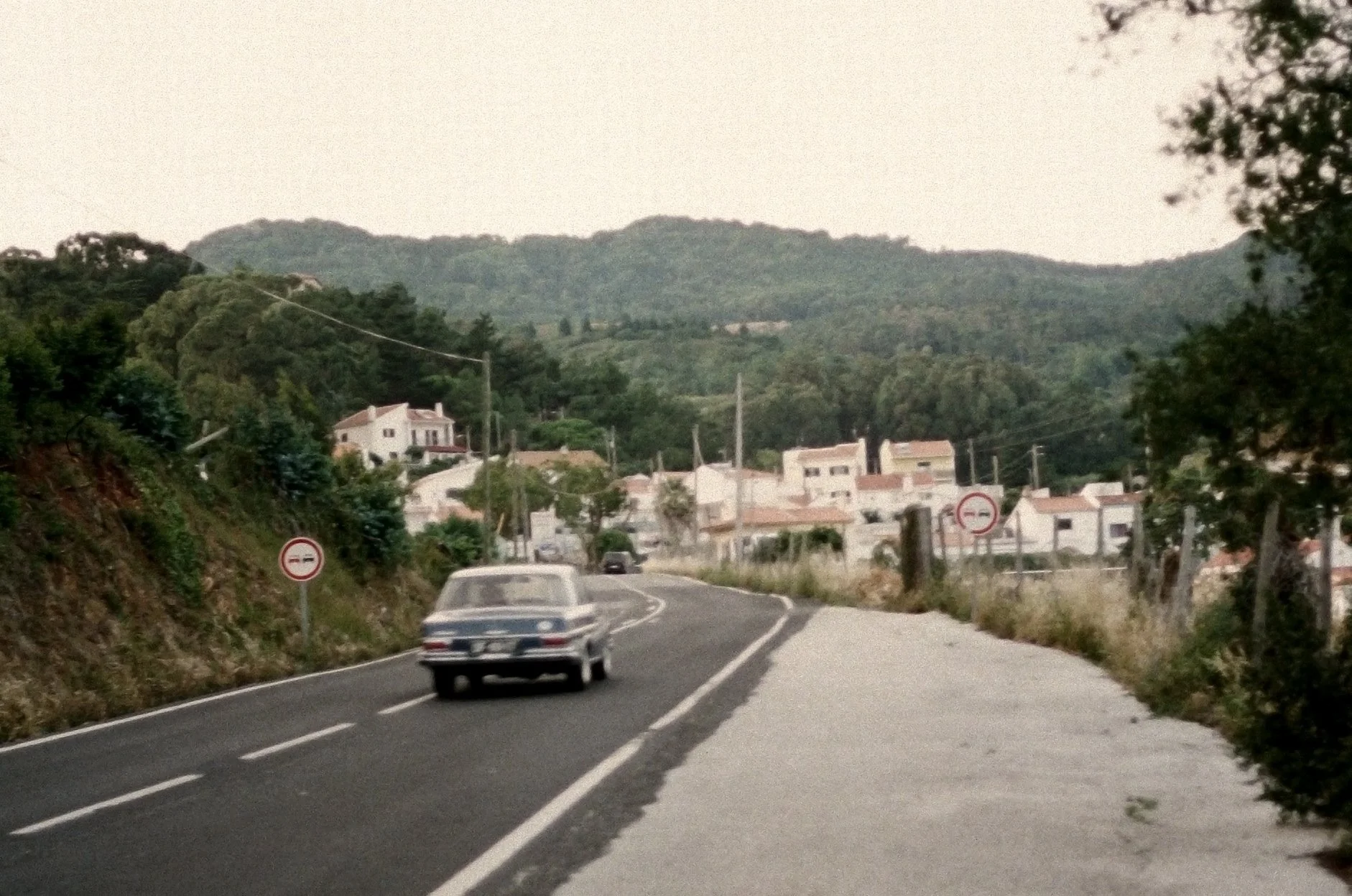 A vintage car driving on a winding country road with houses and green hills in the background.