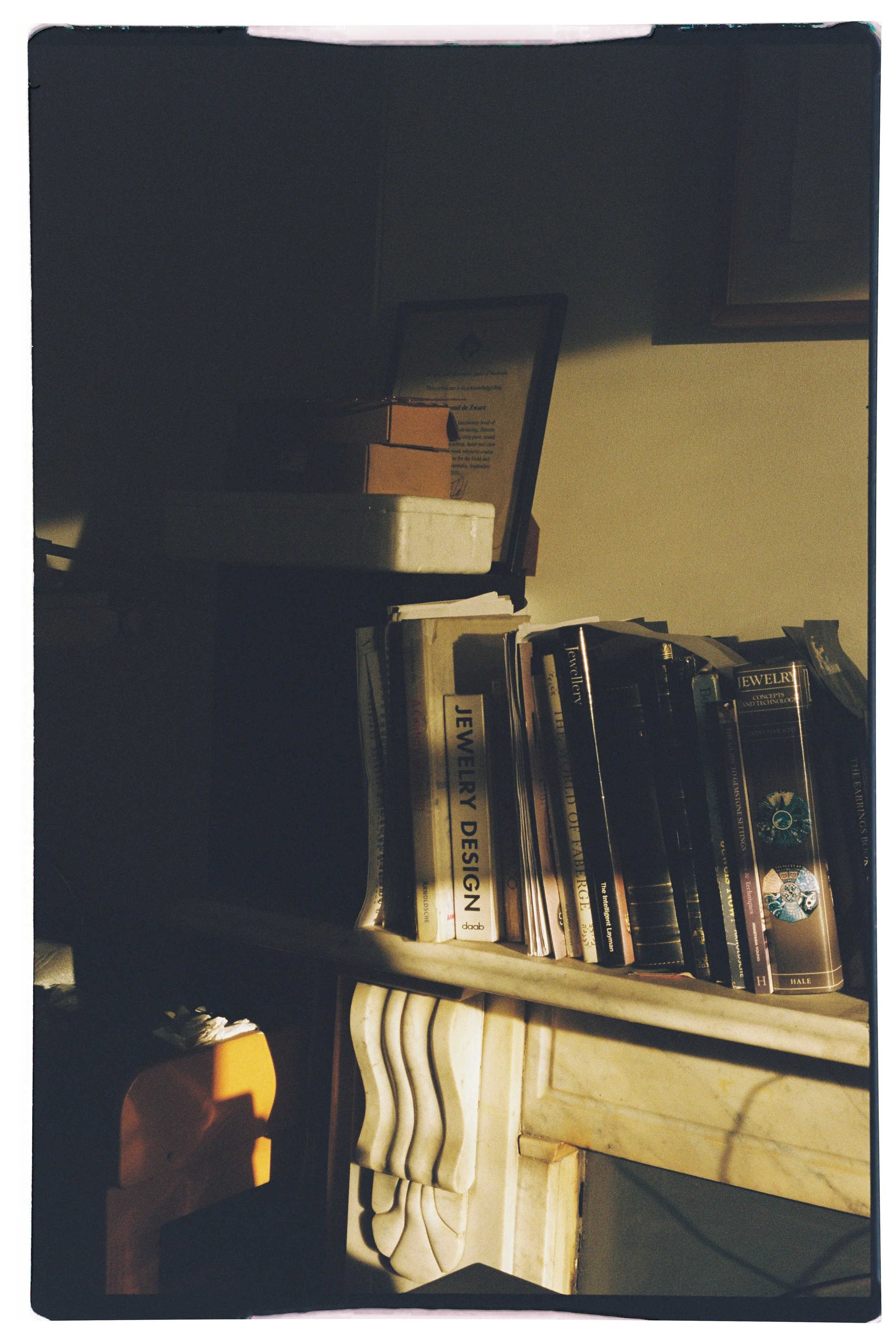 A shelf filled with books on jewelry design, with sunlight casting shadows on the books and a marble surface underneath.
