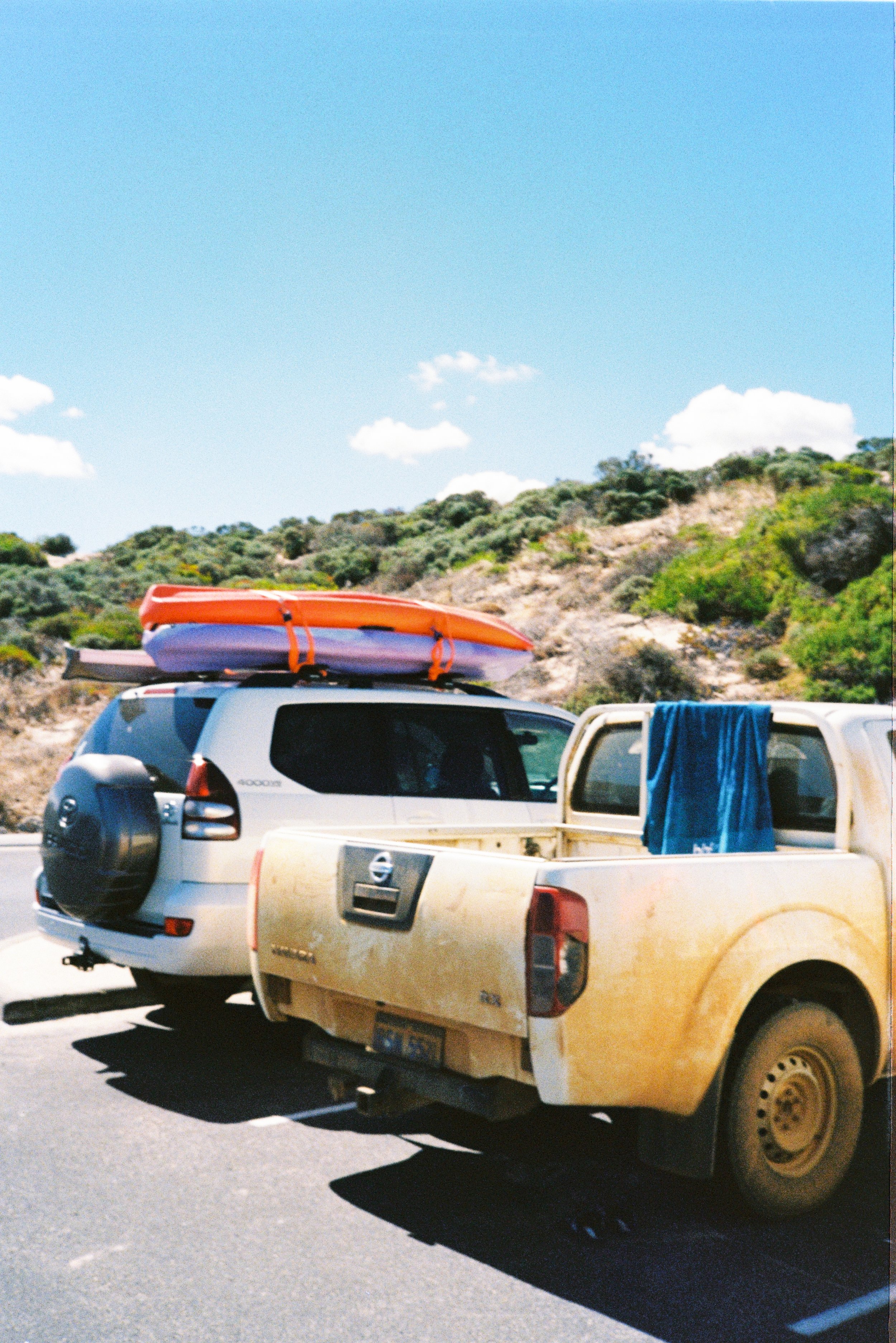 A white SUV with two kayaks on its roof parked next to a yellow pickup truck with a towel hanging on its window in a parking lot near a hilly, green landscape under a bright blue sky with a few clouds.