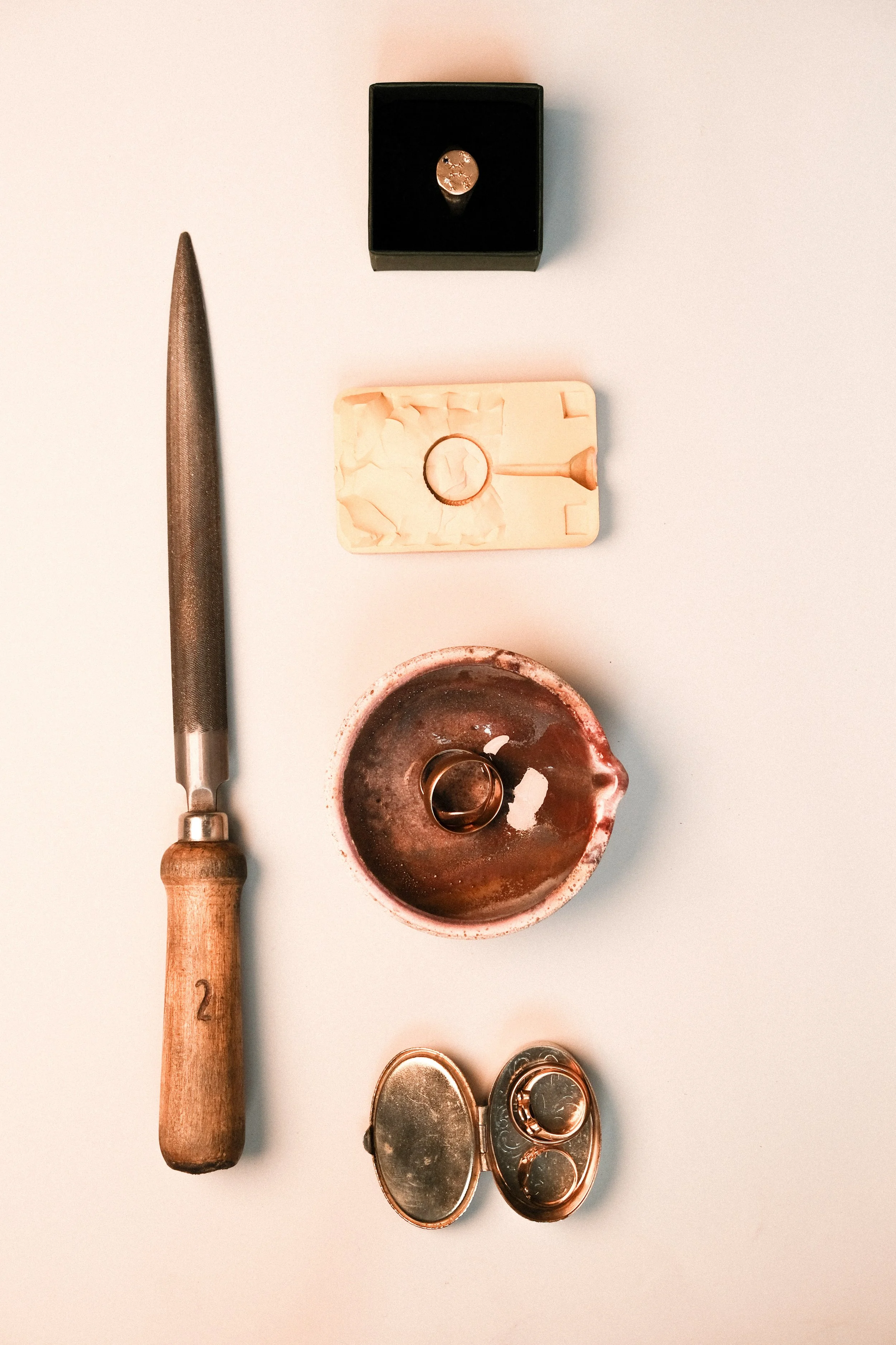 Jewelry ring making tools, including a handmade ring, in a pink ceramic dish, and small ring molds and a ring box on a white surface.