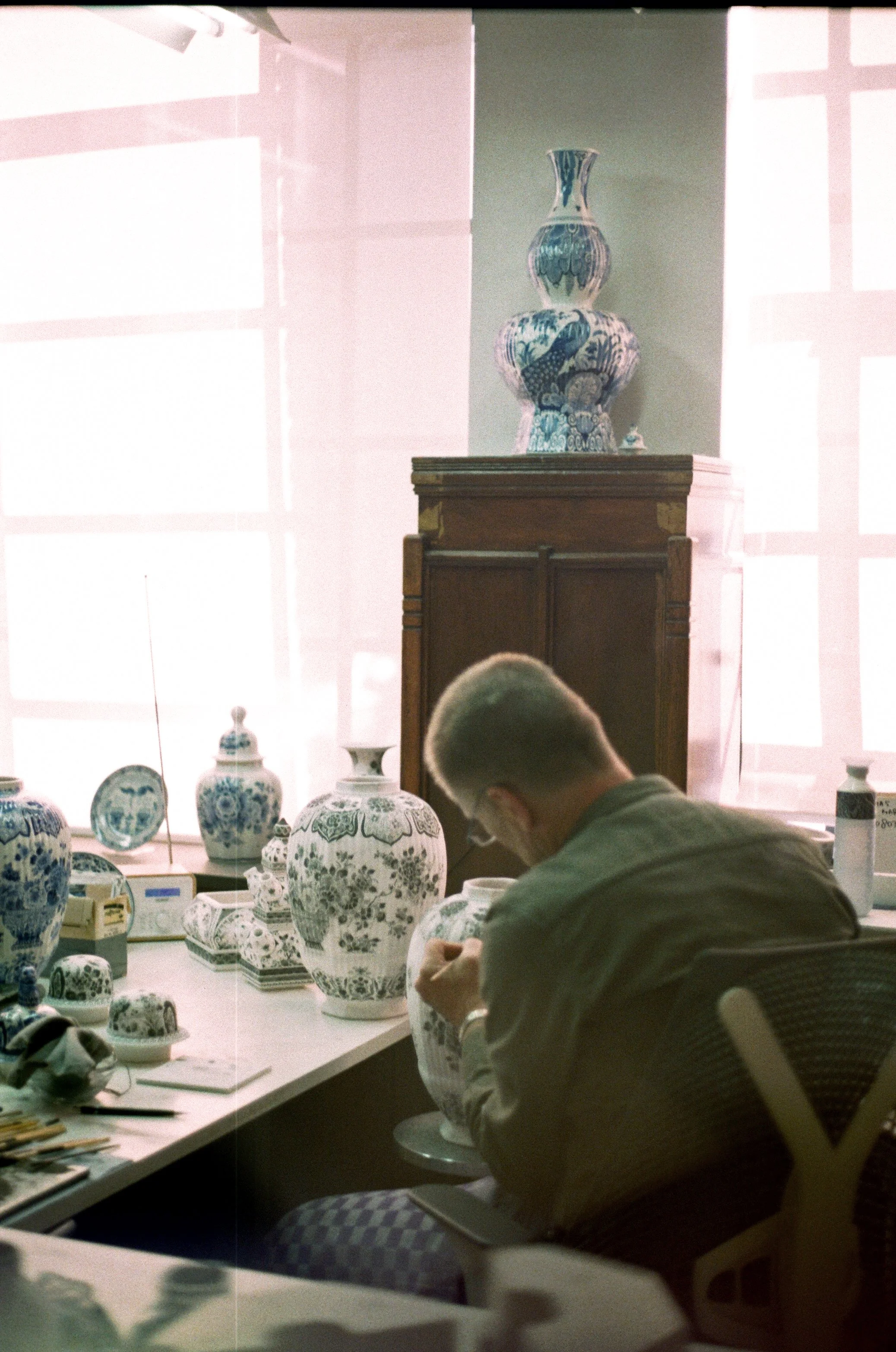 A person sitting at a table examining a large decorative ceramic vase with a floral and figurative pattern, surrounded by various other ceramic pieces and tools, in a well-lit room with large windows and a display of ceramic vases on a wooden cabinet.