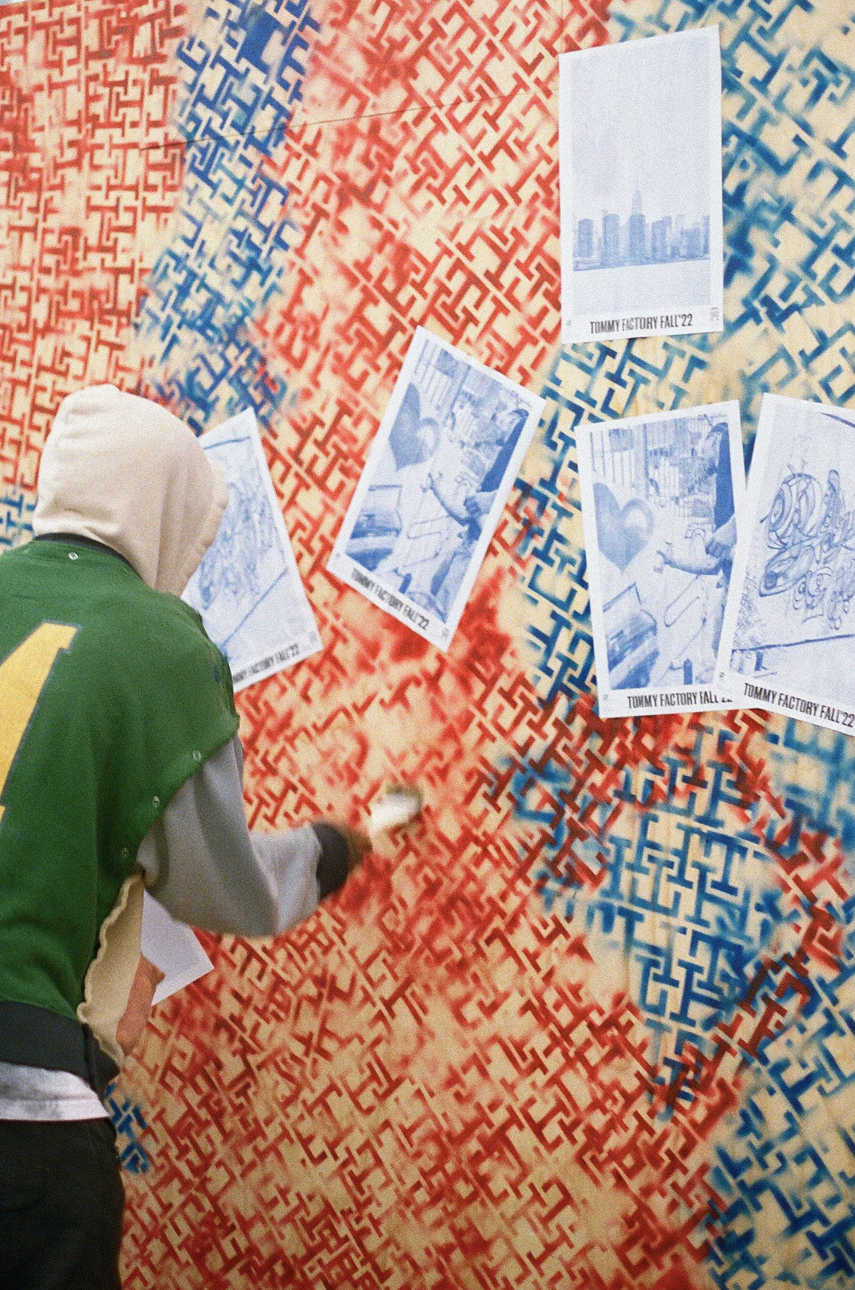 A person in a green and gray jacket with a beige hoodie, spray painting a red and blue geometric mural on a wall with black spray paint. Several blue and white photographs are taped to the wall, showing scenes from a production titled 'TOMMY FACTORY FALL 22'.