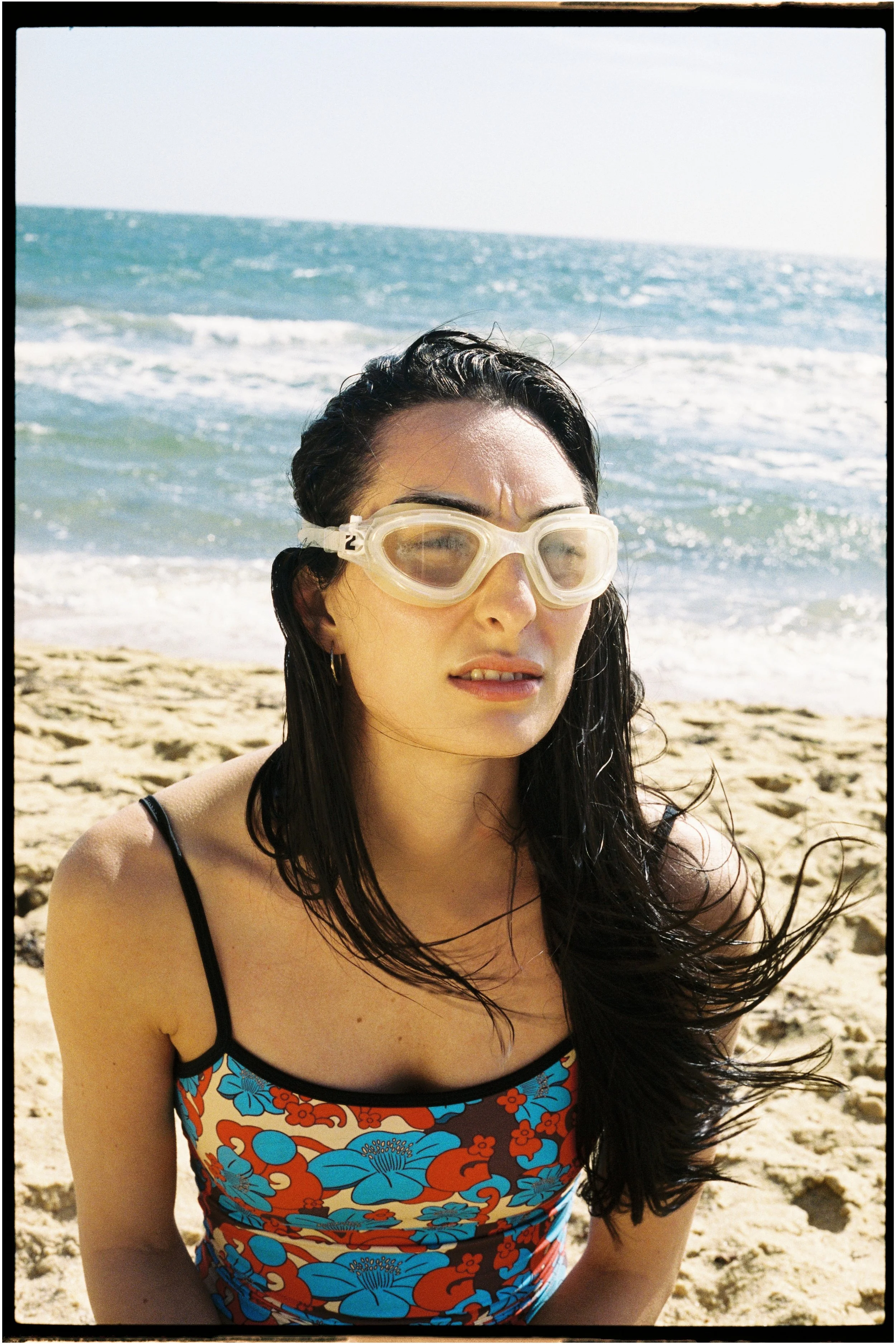 A woman with dark hair wearing goggles and a colorful floral swimsuit sitting on a sandy beach with waves and ocean in the background.