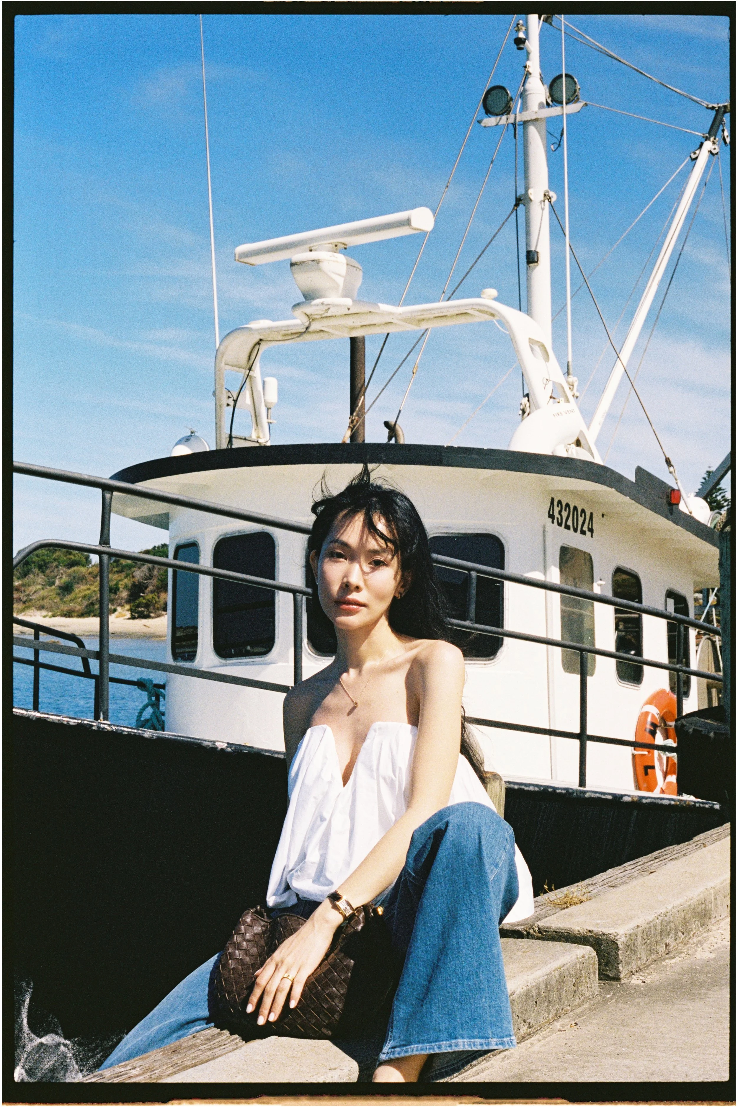 A woman sitting on a dock in front of a boat, with a blue sky and water in the background.