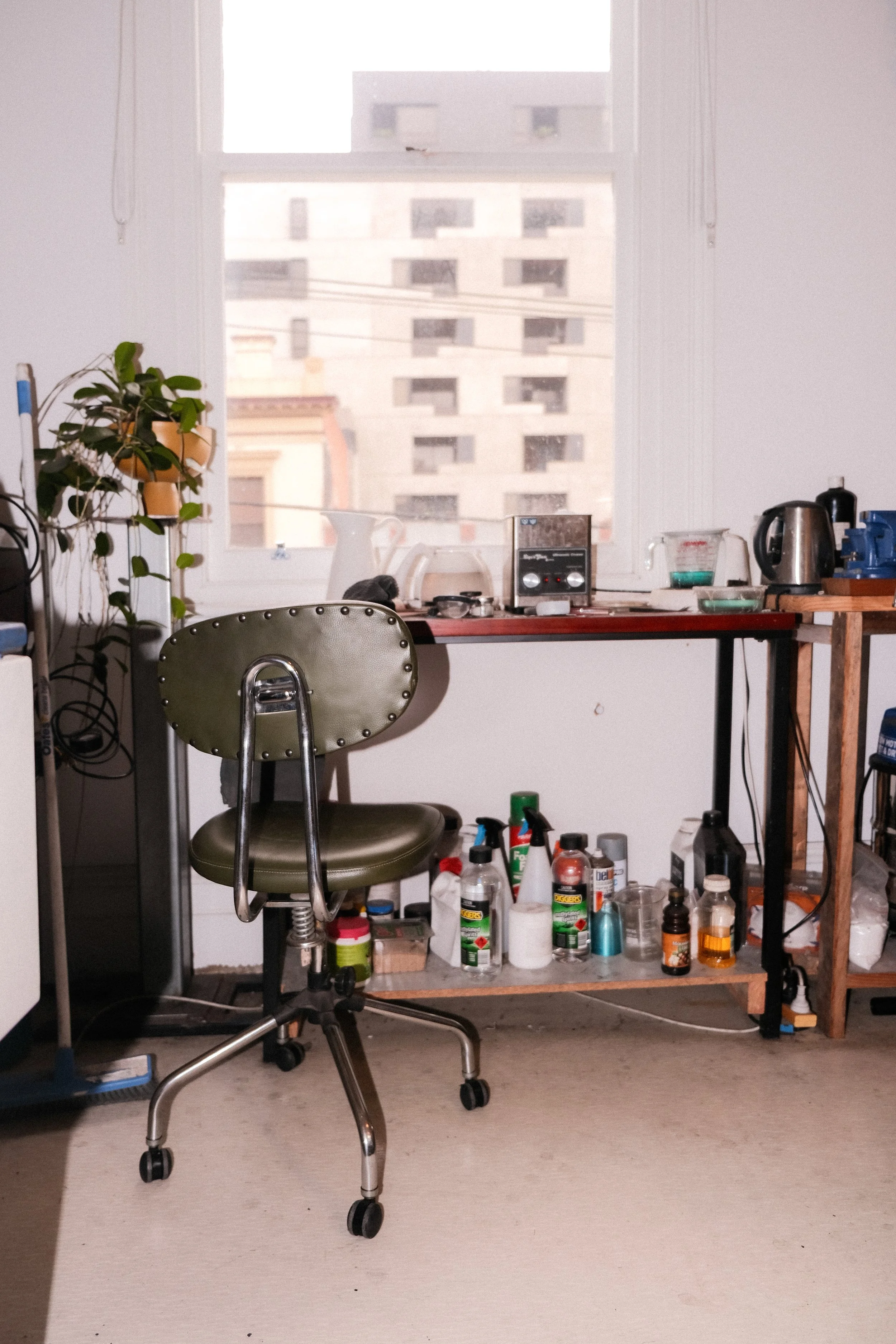 A cluttered workspace with a green office chair, a wooden desk with various items including a kettle, a measuring cup, and a stereo, and shelves below with cleaning supplies and bottles. There is a large window with a city view in the background.