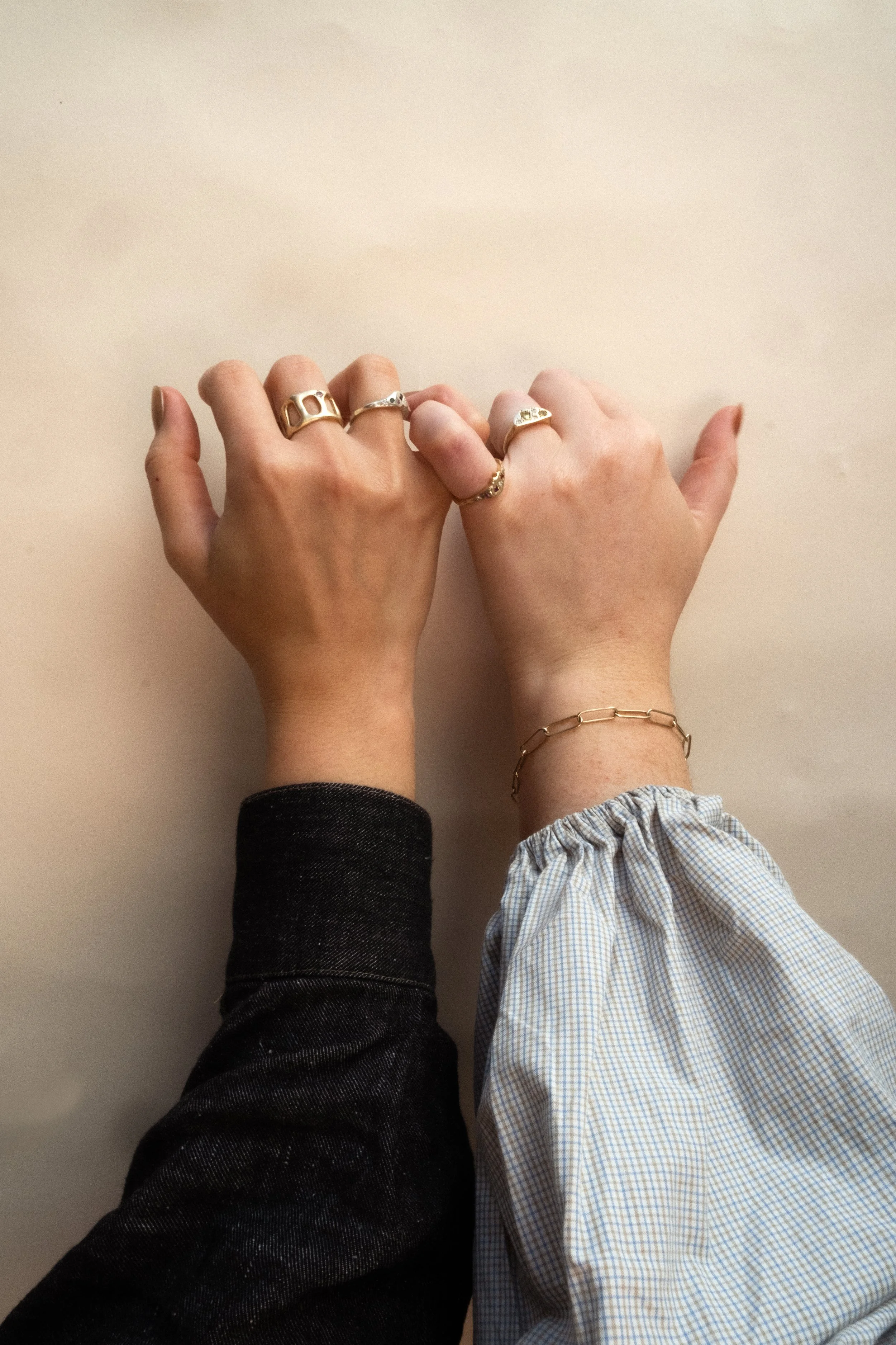 Two hands with jewelry resting on a light-colored surface, one with a dark sleeve and the other with a checkered sleeve.