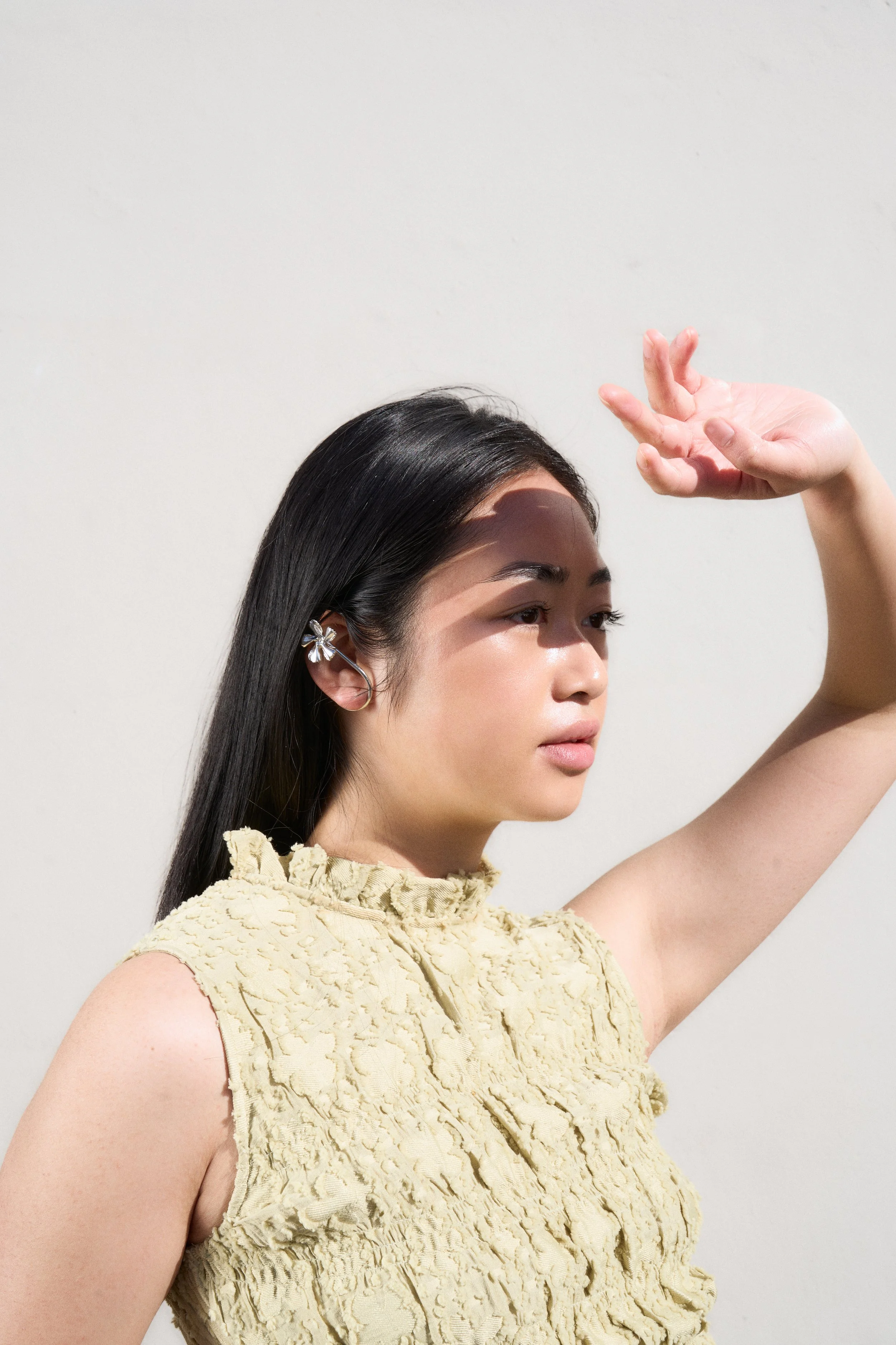 A young woman with long black hair, wearing a beige ruffled sleeveless top, is raising her right arm with her hand near her forehead, looking into the distance against a plain white wall.