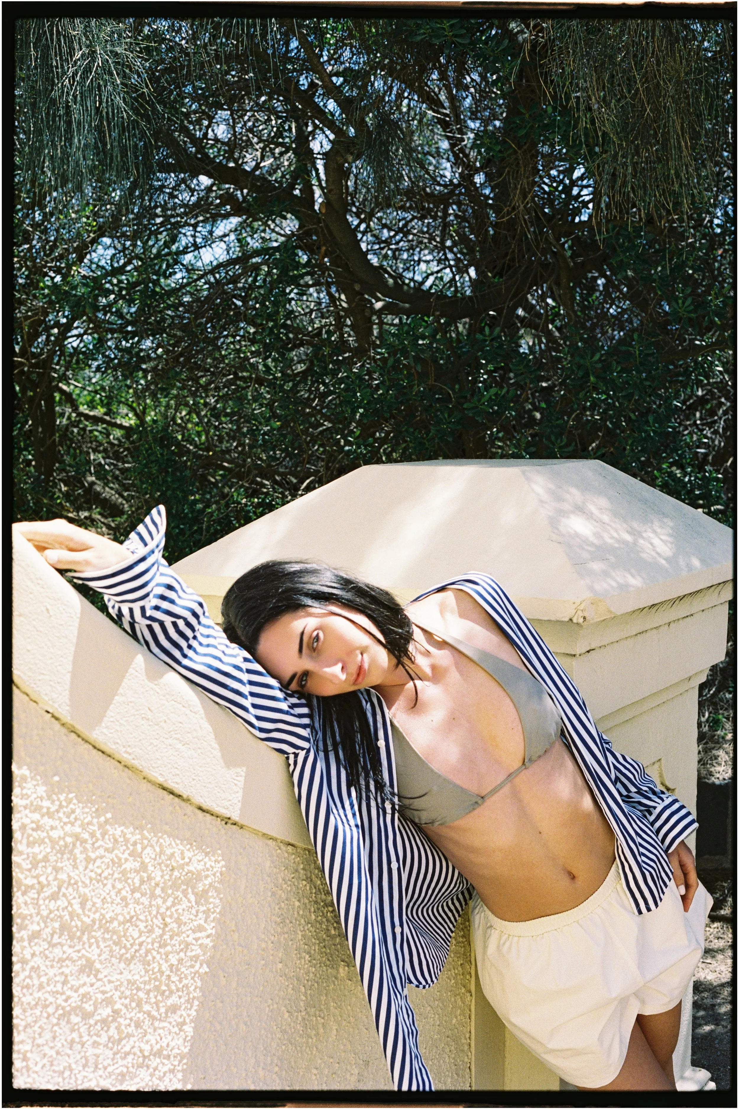 A woman leaning against a beige wall outdoors, wearing a striped shirt, bikini top, and shorts, with trees and blue sky in the background.