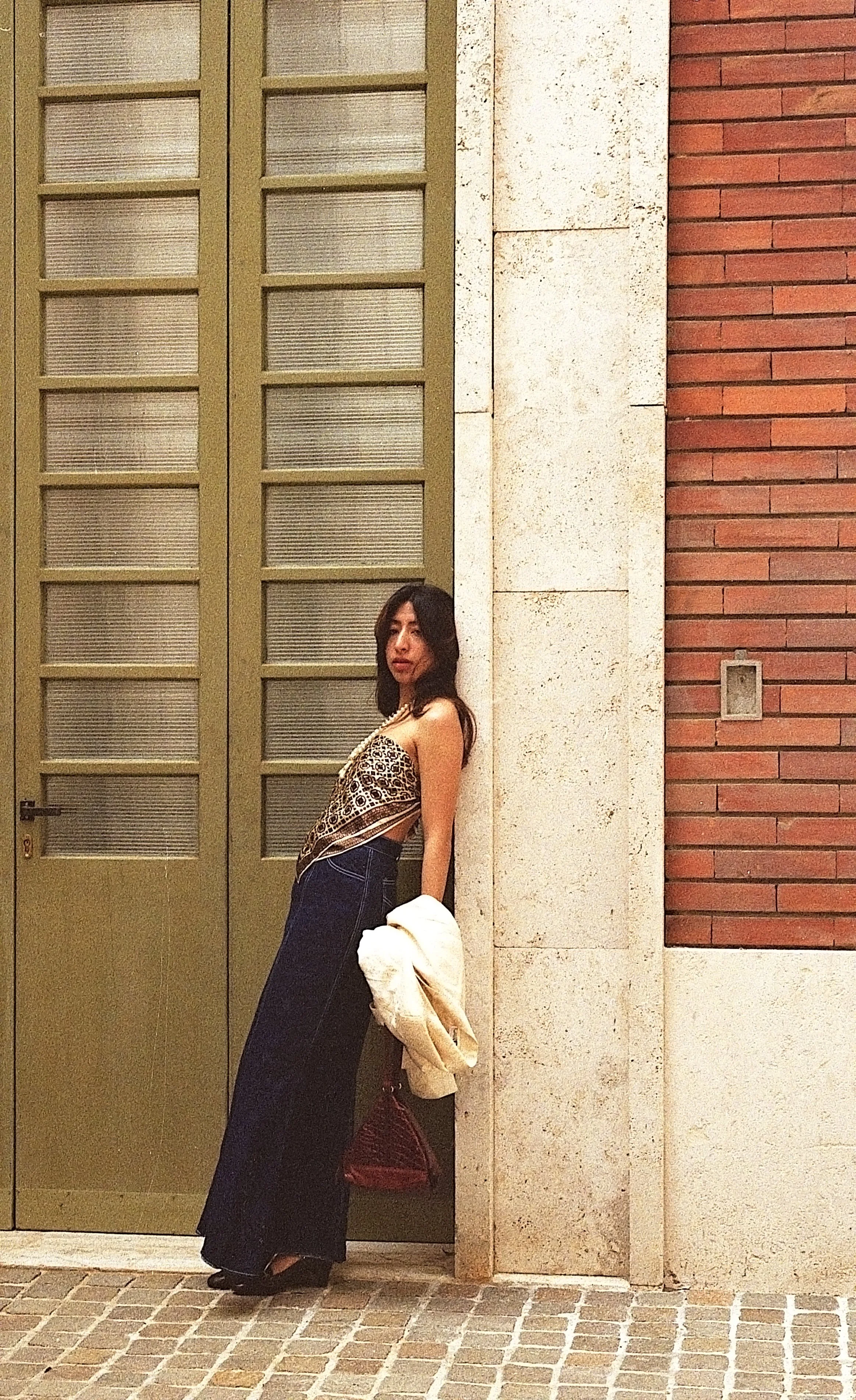 A woman leaning against a wall outside a building, holding a jacket and a patterned purse. She is wearing a leopard-print top and dark wide-leg jeans.