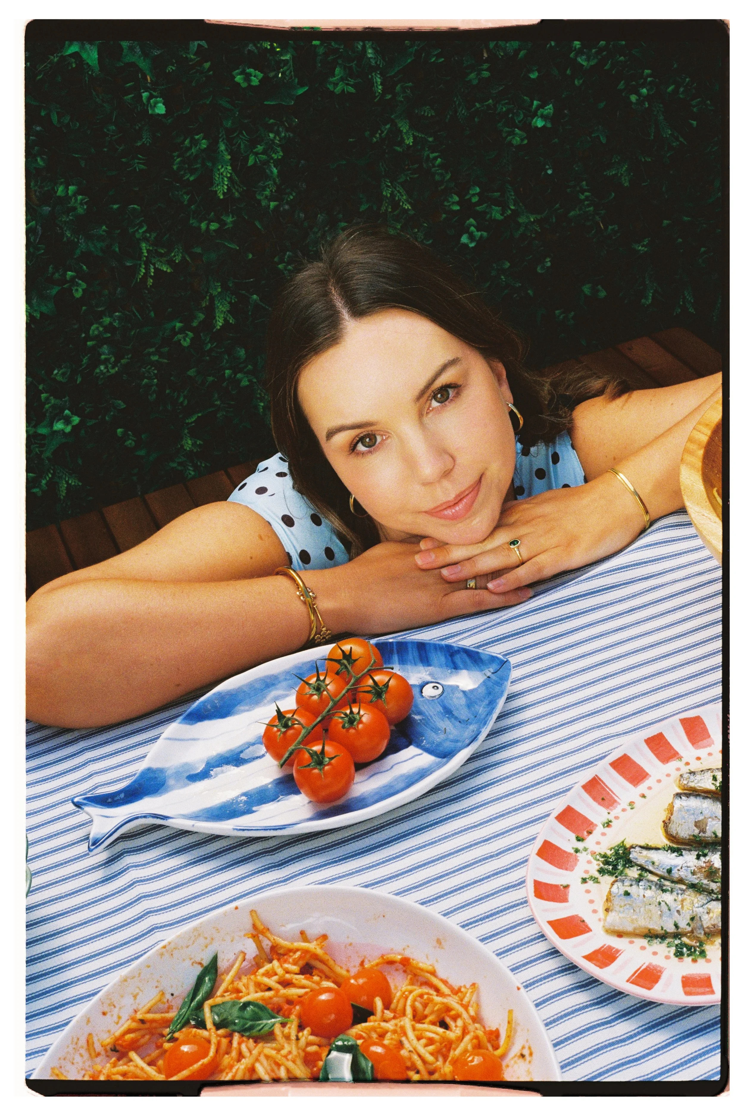 A woman lying at a table with food, resting her head on her hands and looking at the camera. There are cherry tomatoes and pasta on the table, with a leafy green backdrop.