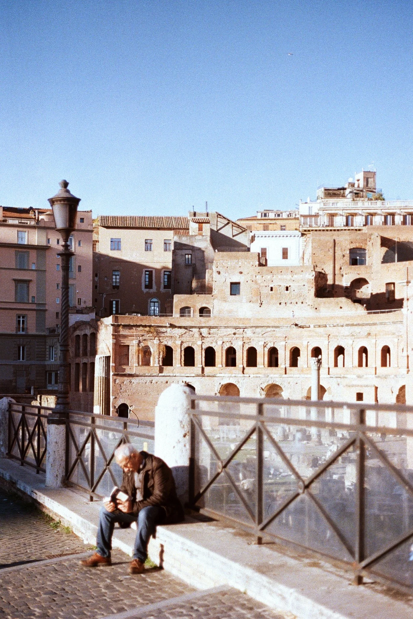 An older man sitting on a bench by a railing, reading a book with ancient ruins and buildings in the background under a clear blue sky.