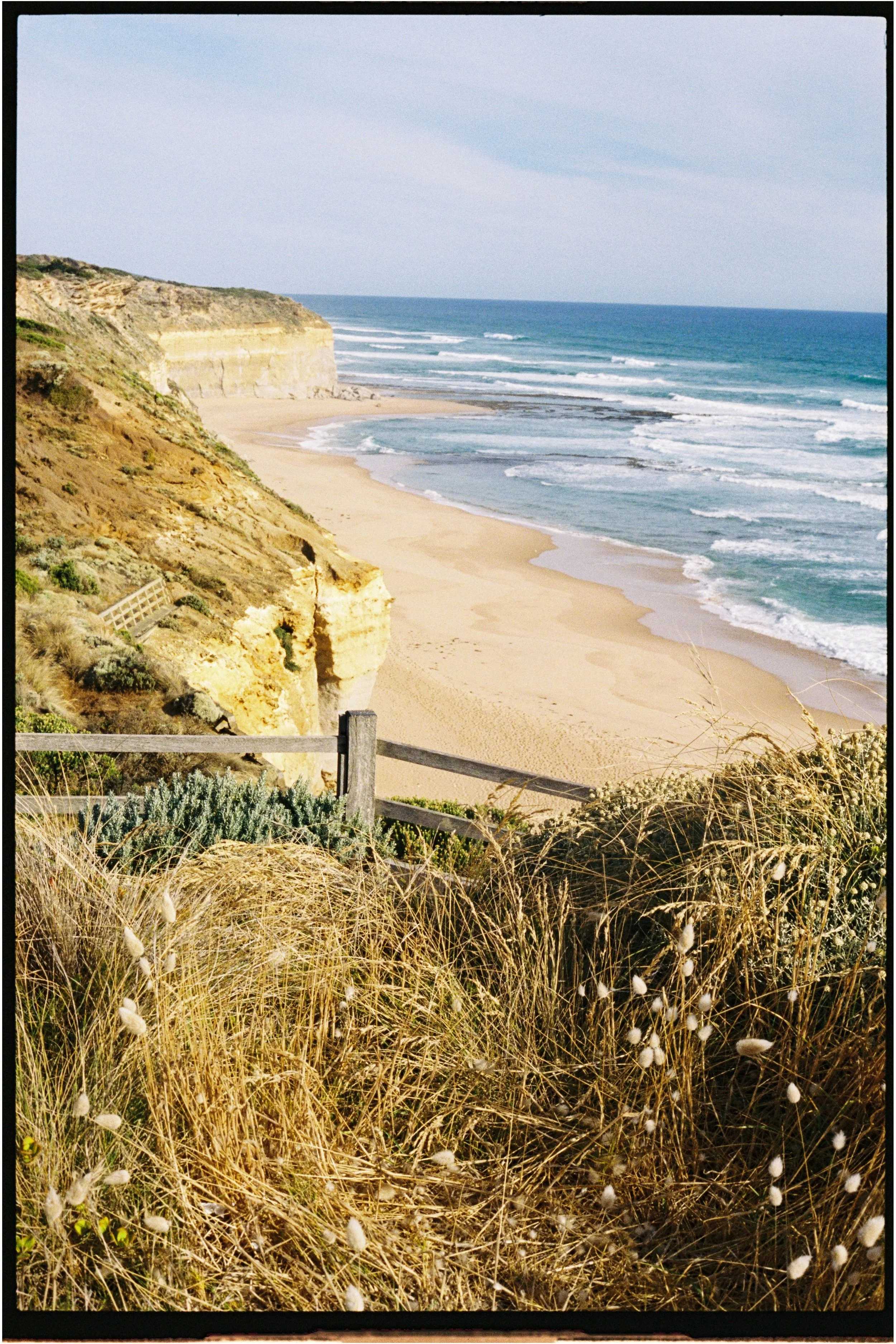 Coastal scene with sandy beach, ocean waves, and cliffs in the distance, with dry grass in the foreground.