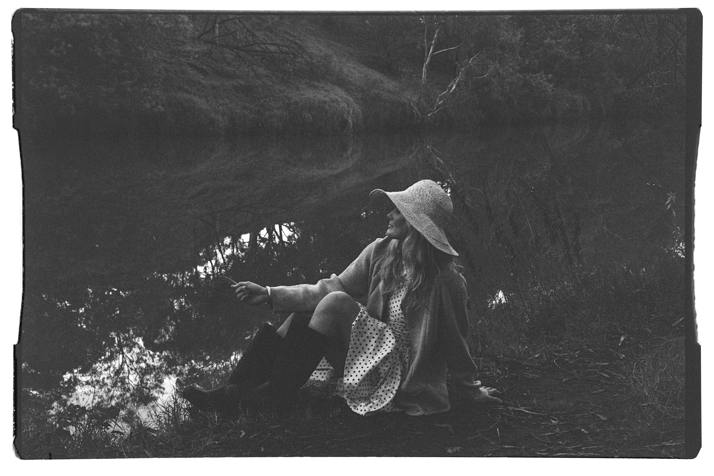 A woman sitting on the ground by a river, wearing a wide-brimmed hat, coat, and dress with polka dots, holding a flower in her hand. The scene is in black and white with trees reflected in the water.