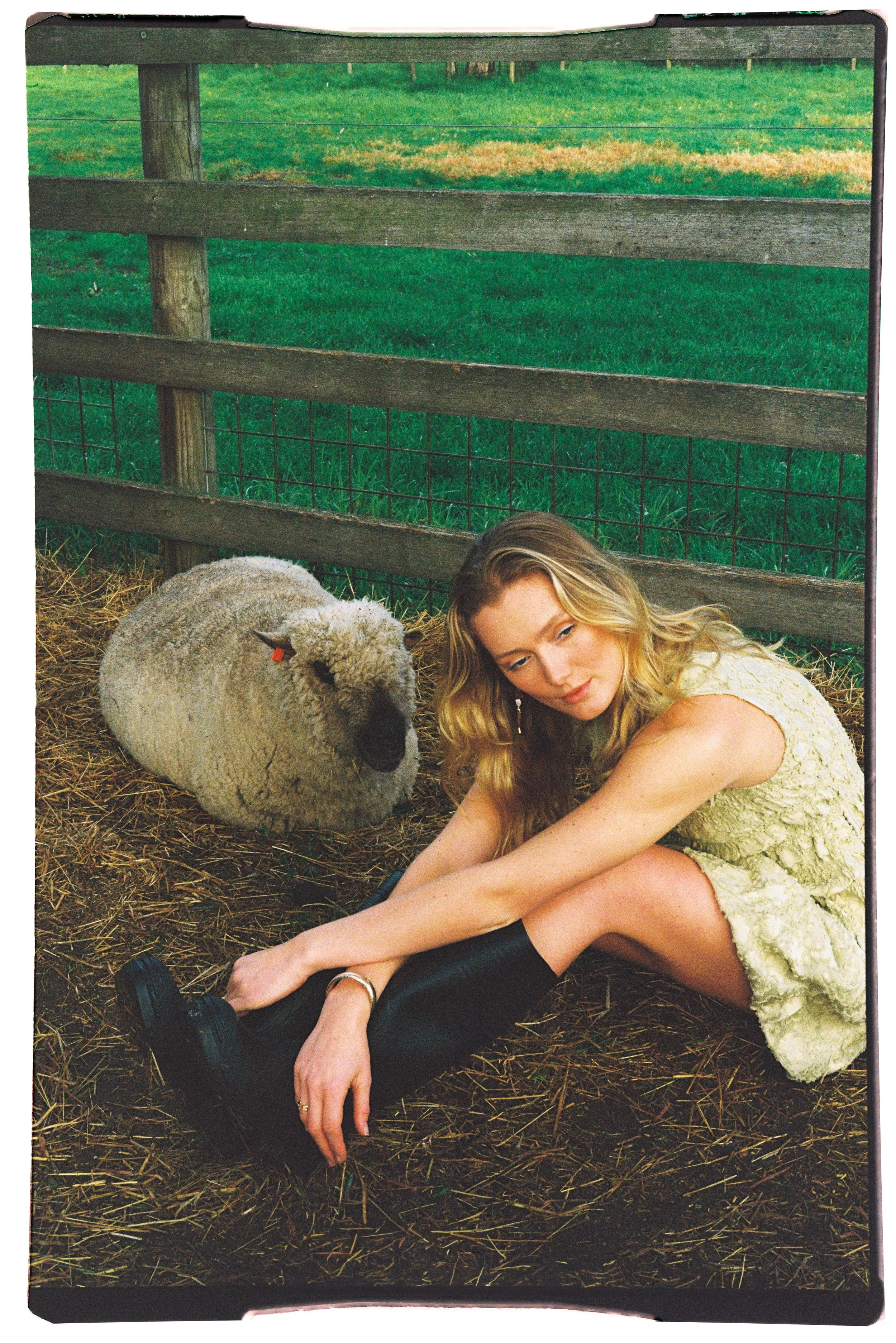 A woman with long blonde hair sitting on the ground next to a sheep in an outdoor barn area with green grass and wooden fencing in the background.
