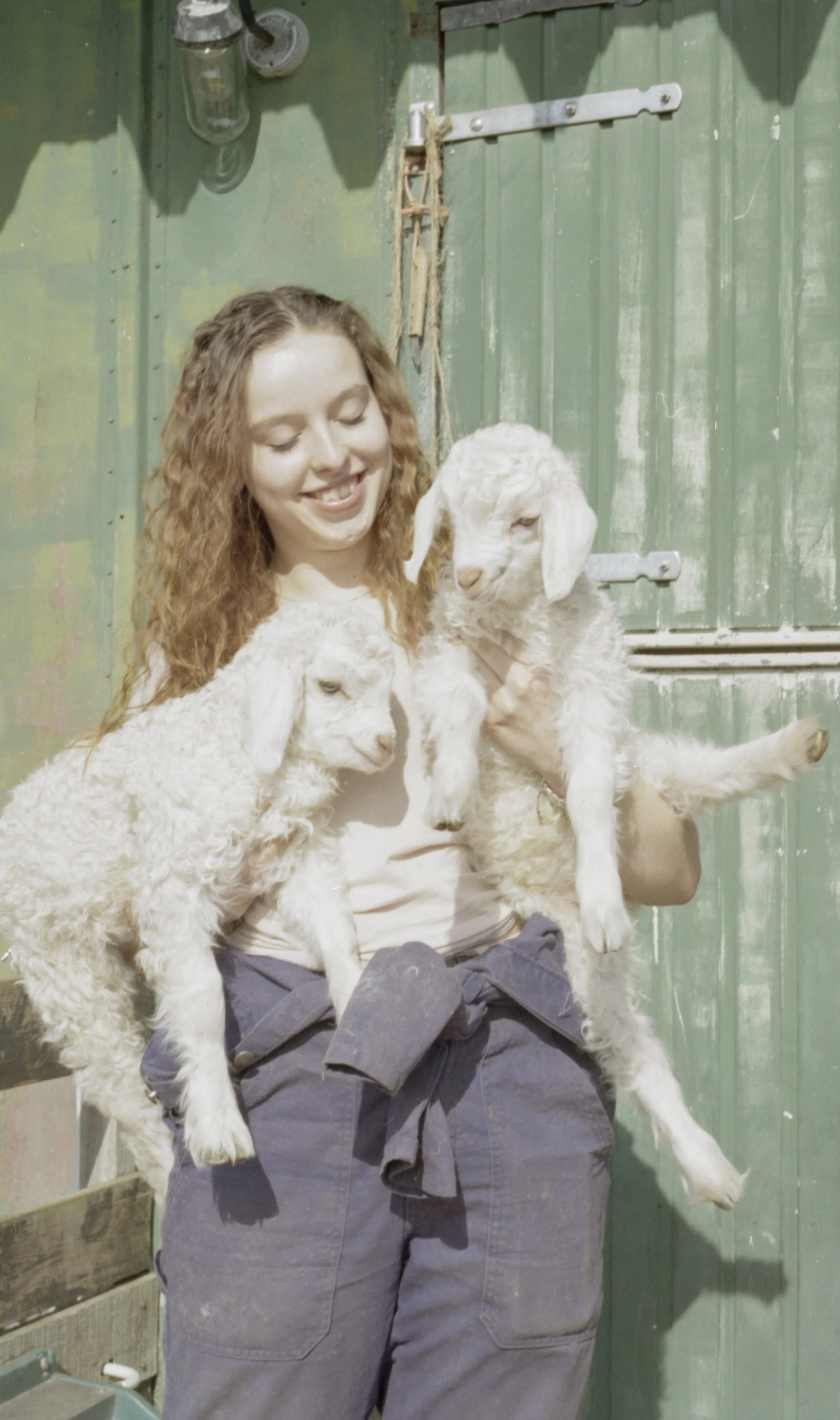 A young woman with curly red hair smiling and holding two white baby goats, one in each arm, in front of a green barn door.