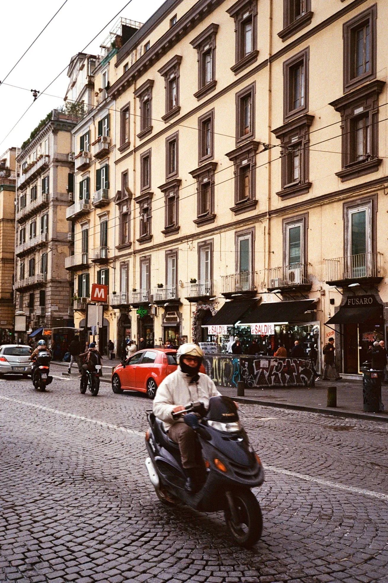 Street scene with a person riding a scooter in the foreground, cars parked along the cobblestone street, and multi-story buildings with balconies and various storefronts in the background.