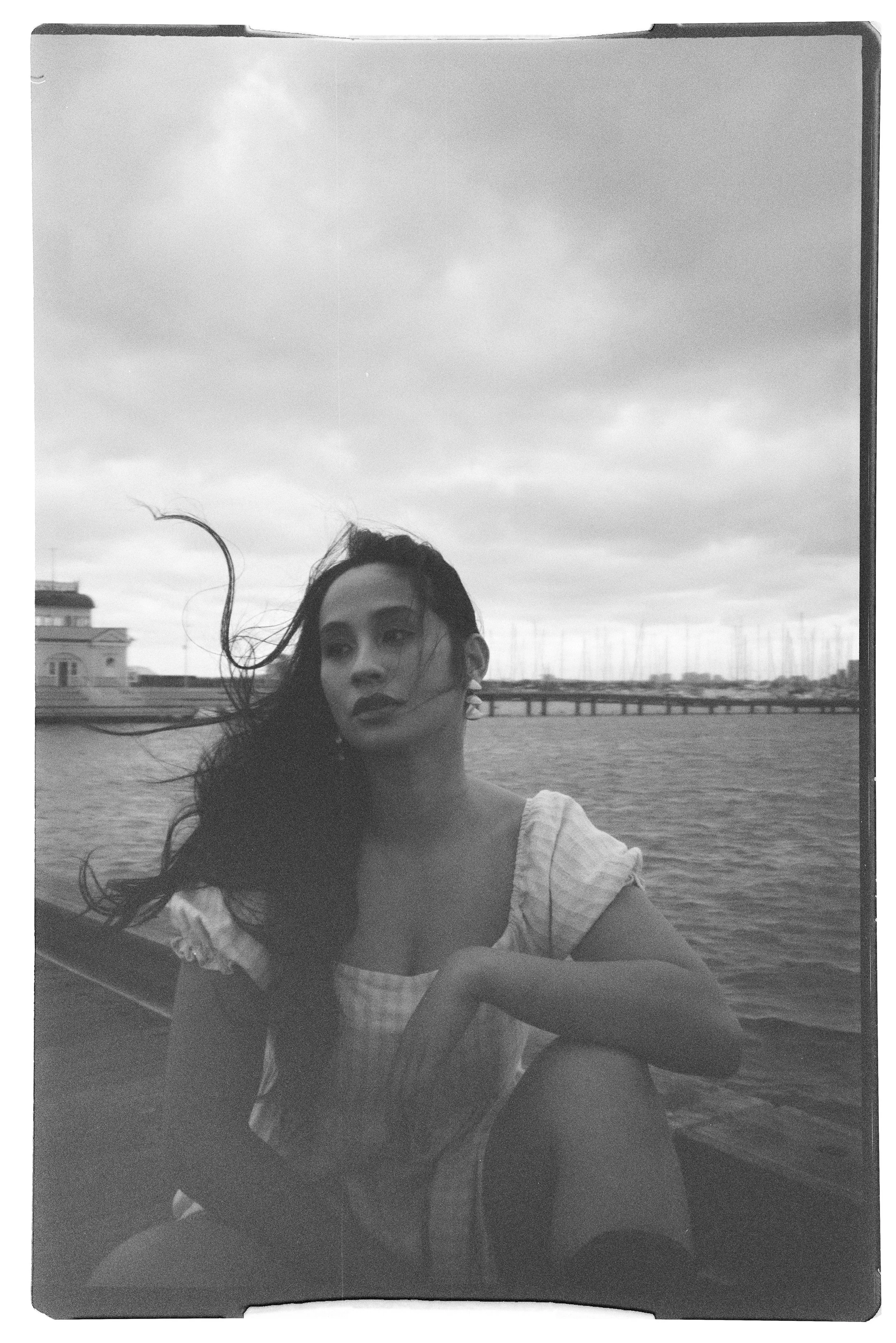 A black and white photo of a woman with long dark hair sitting near a body of water, with a bridge and buildings in the background, under a cloudy sky.