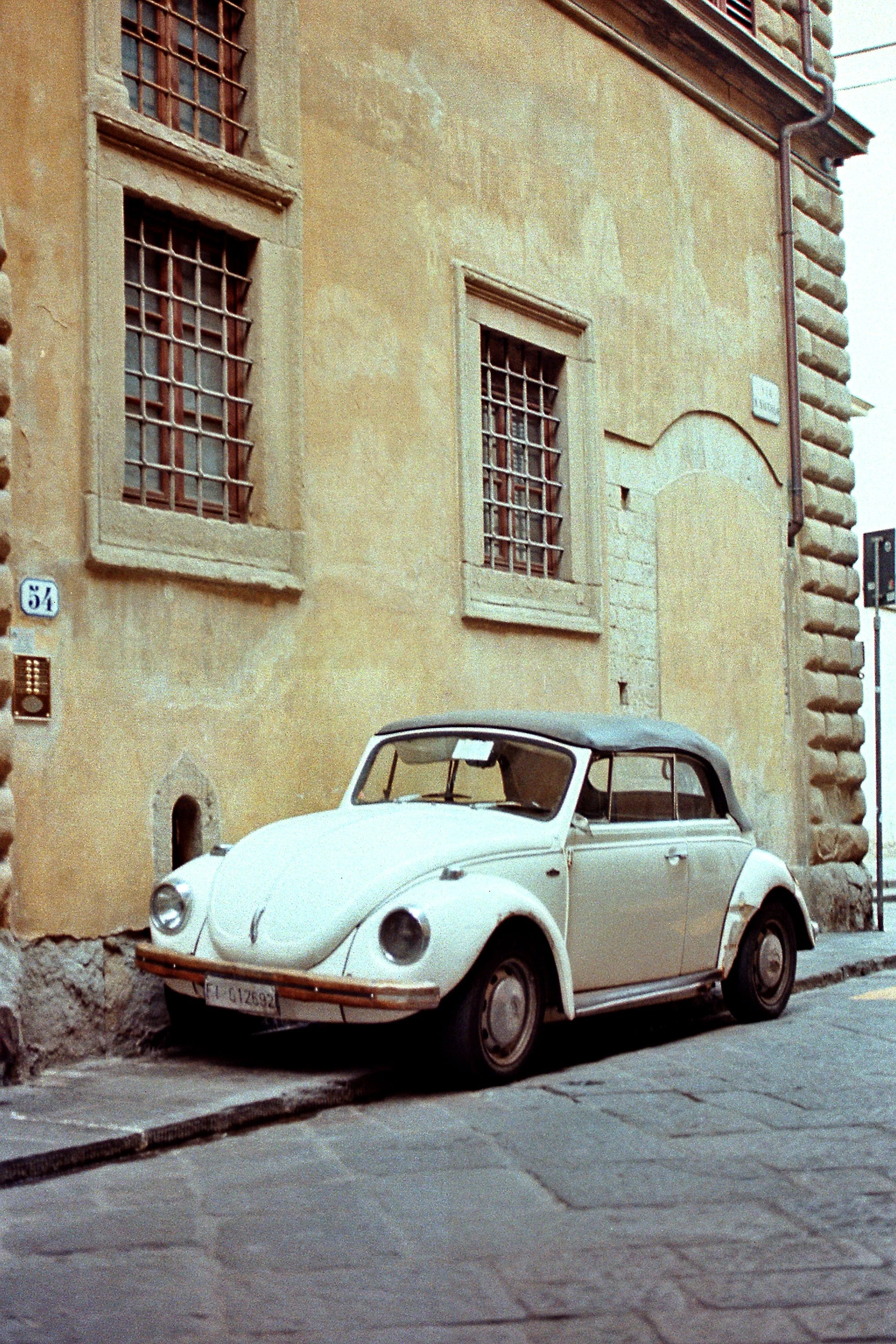 A vintage white Volkswagen Beetle parked on a cobblestone street in front of an old yellow stucco building with barred windows and a small plaque.