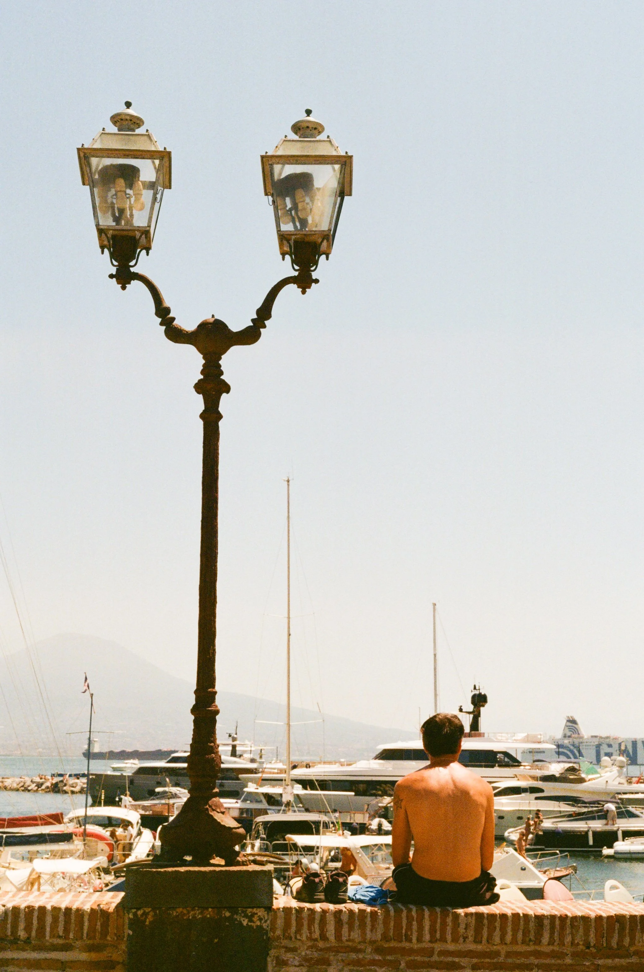 A man sitting on a brick wall by a marina with boats and yachts, near a vintage street lamp, on a hazy day.