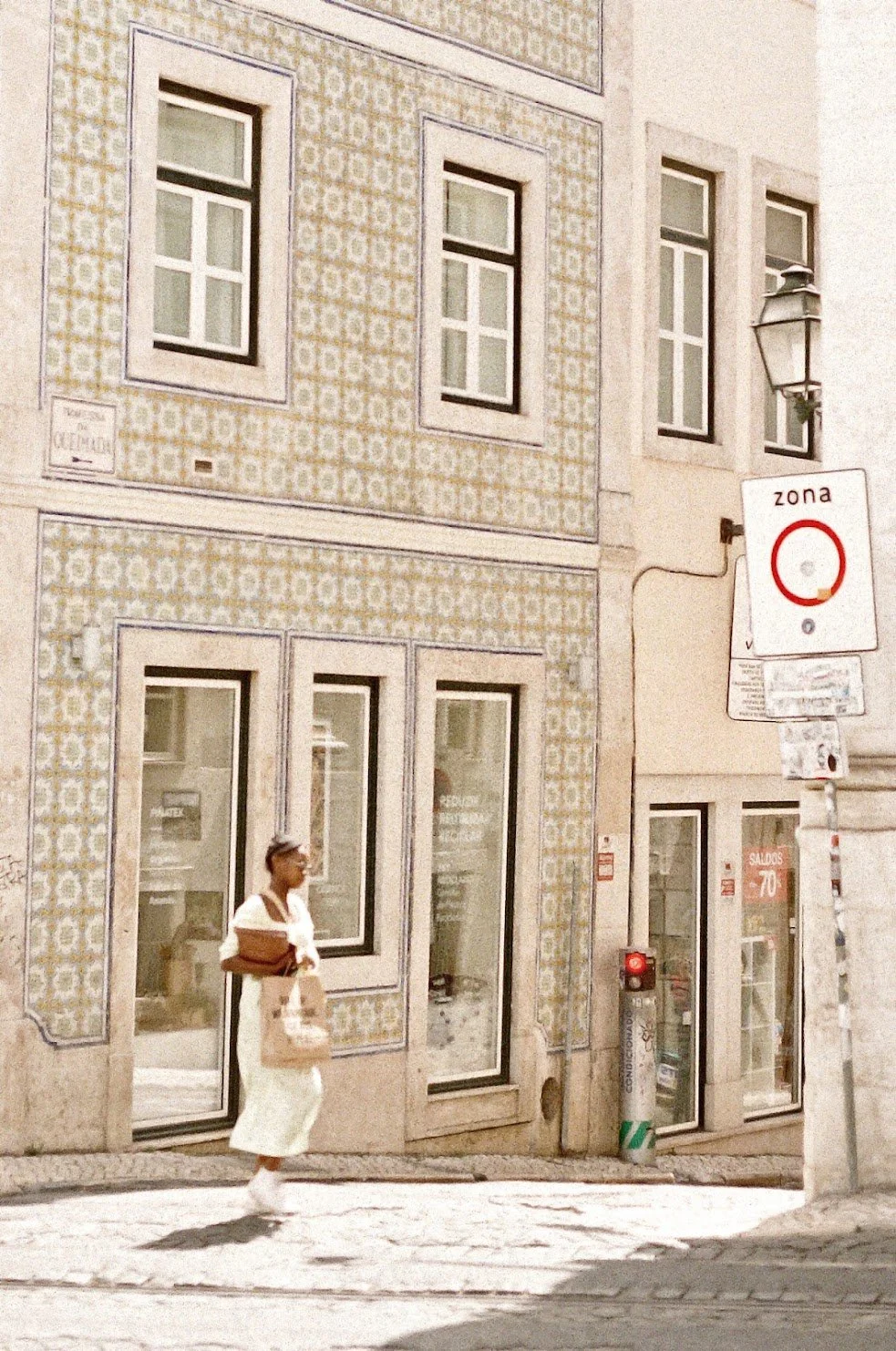 A woman in white with a brown bag crossing the street in front of a building with decorative tiled walls and multiple windows.