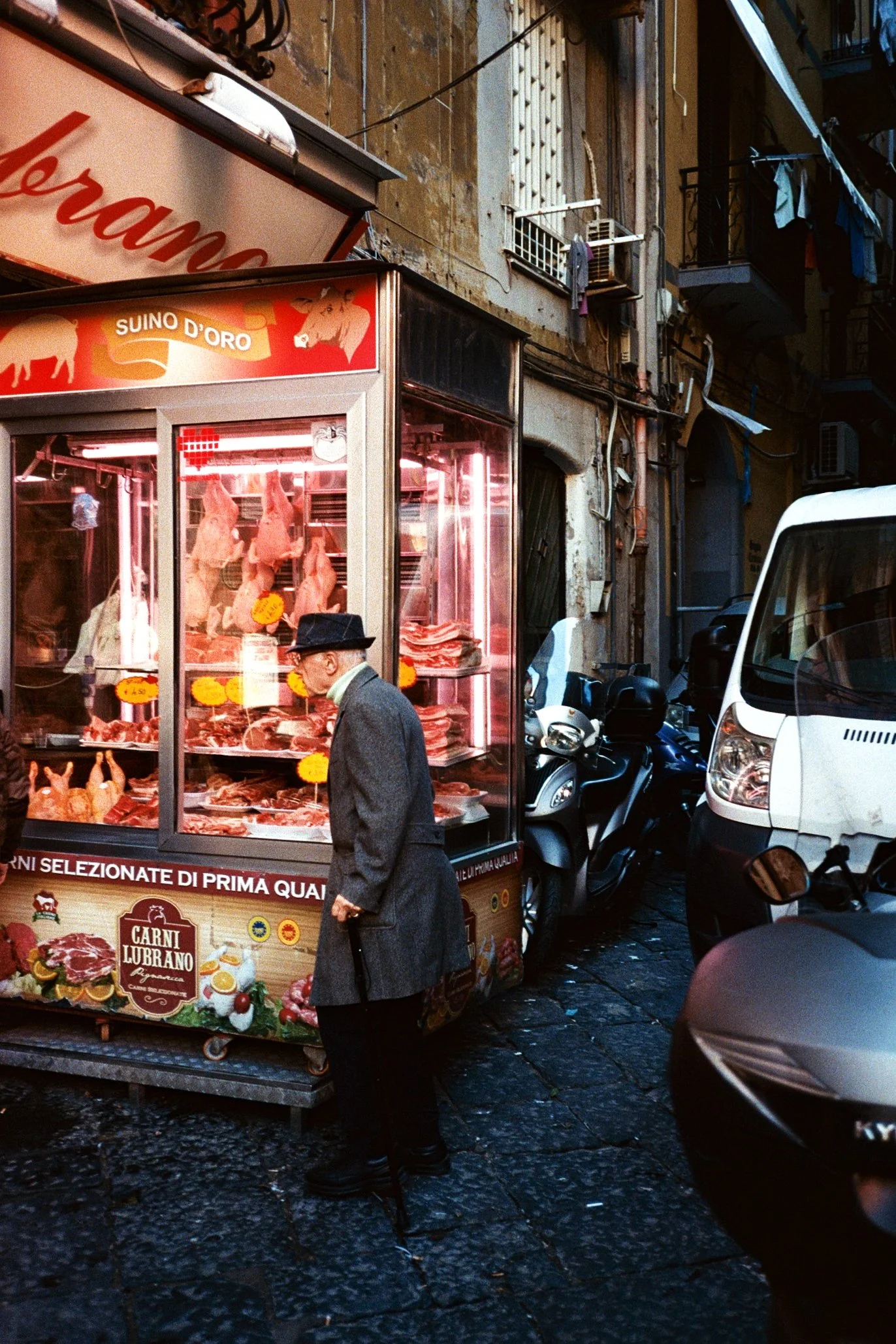 An elderly man in a gray coat and black hat looking at a meat shop on a narrow city street, with parked motorcycles and cars nearby, and old buildings with laundry hanging outside.