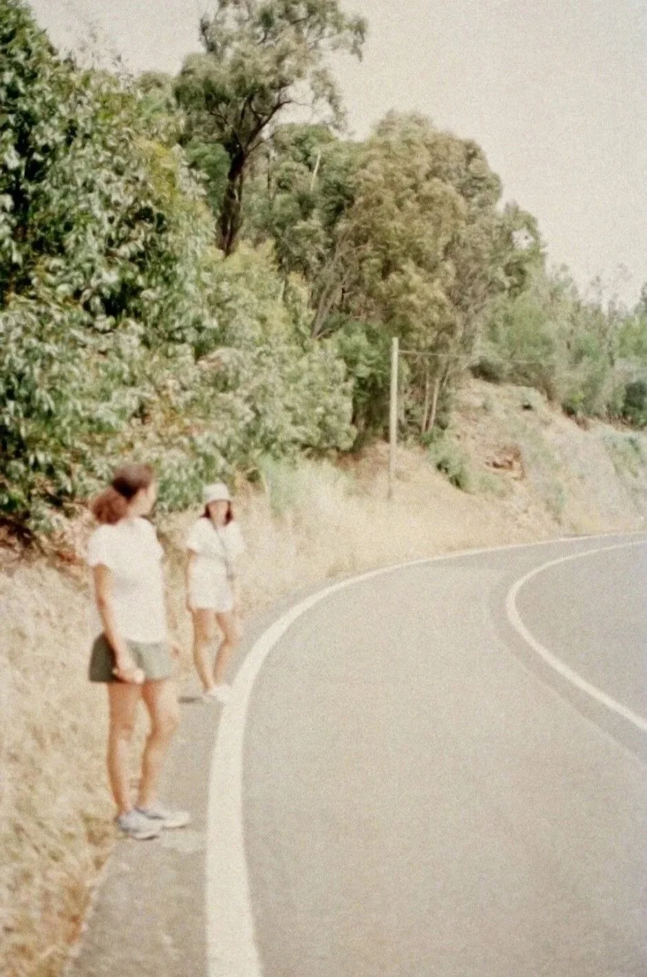 Two women standing on the side of a winding road surrounded by green trees and bushes, with a utility pole in the background.