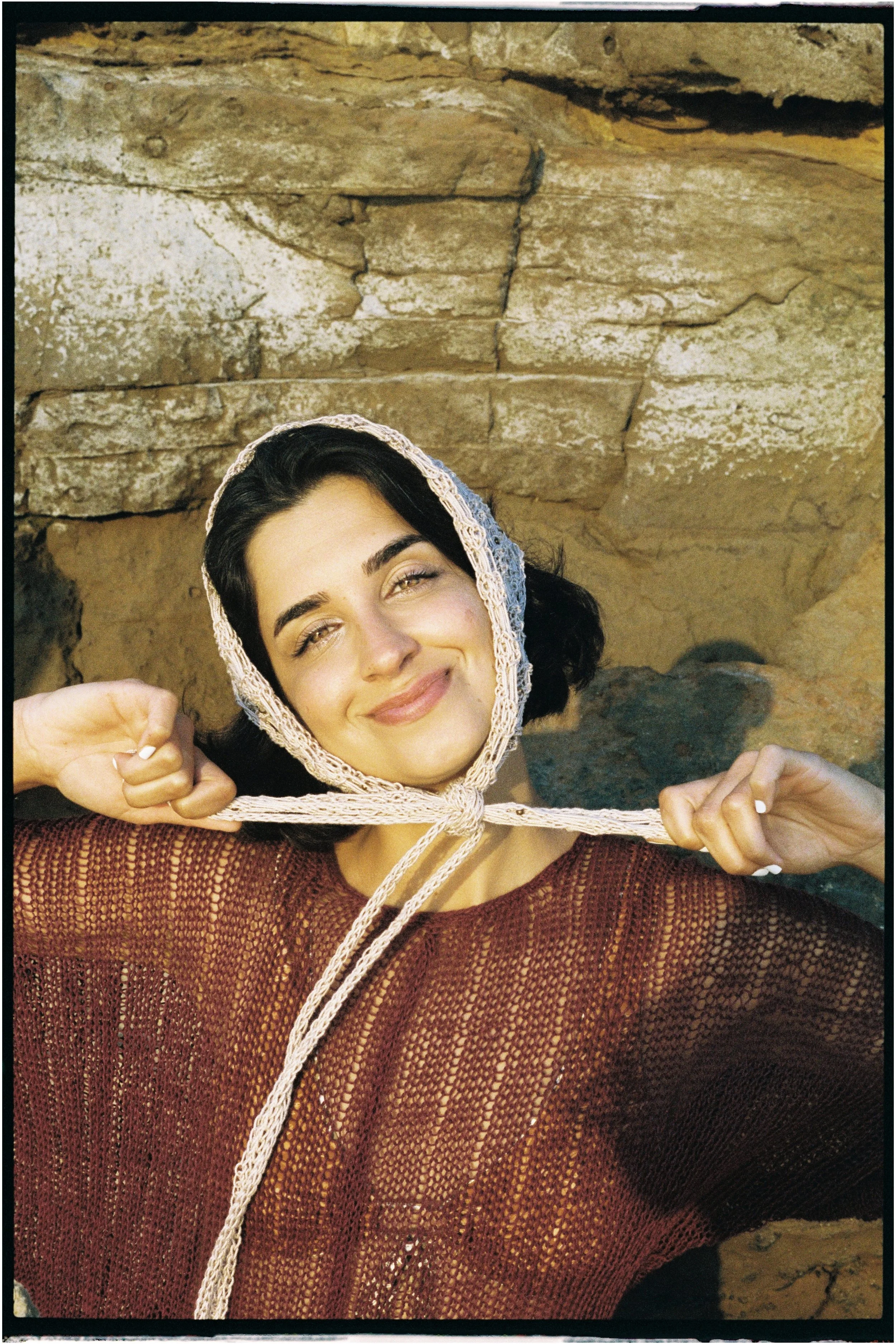 A woman with dark hair, smiling, wearing a beige lace hood and a reddish-brown mesh top, stands against a rocky background and holds the hood with both hands.