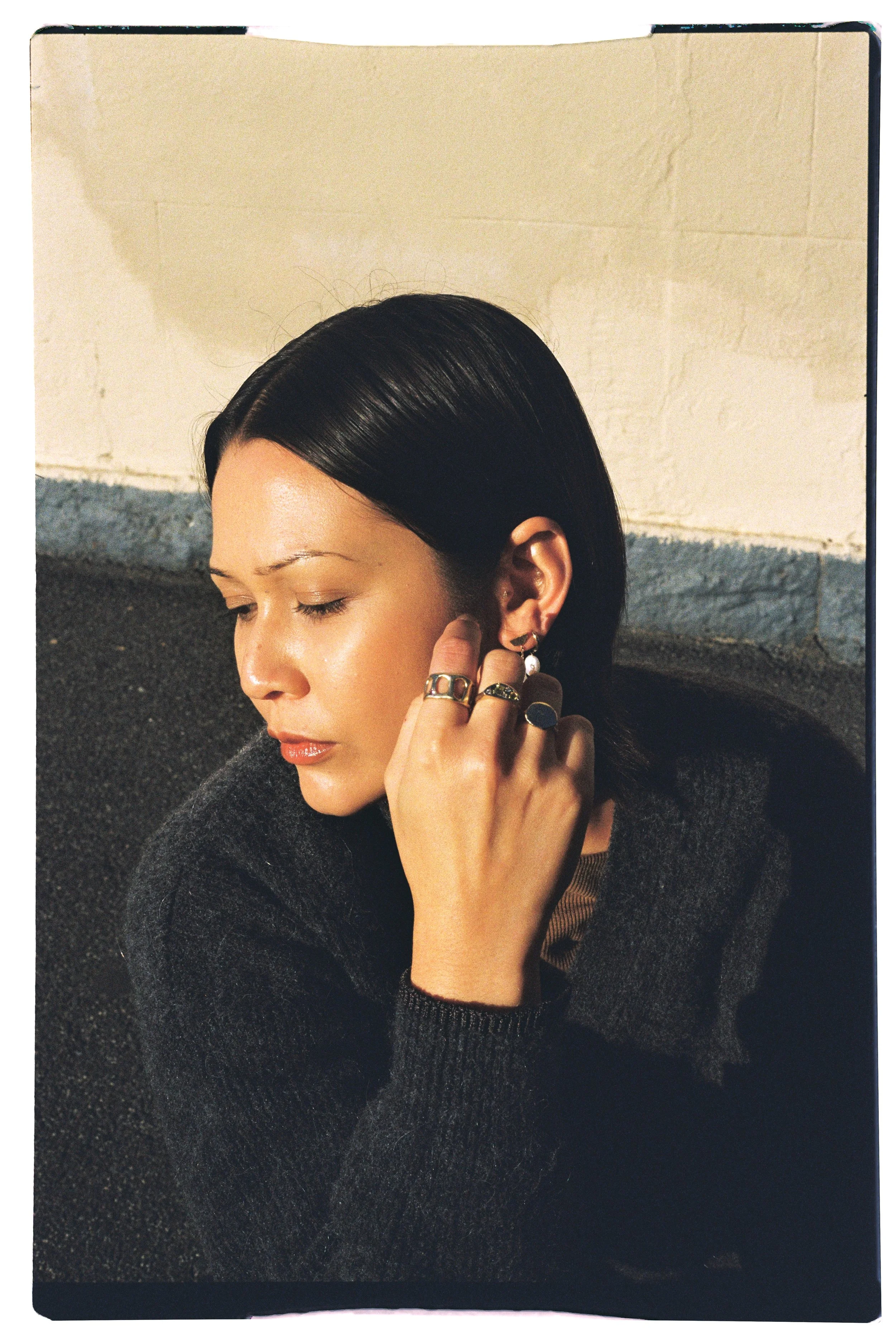 A woman with sleek black hair and jewelry, resting her hand near her ear with her eyes closed, against a beige and gray background.
