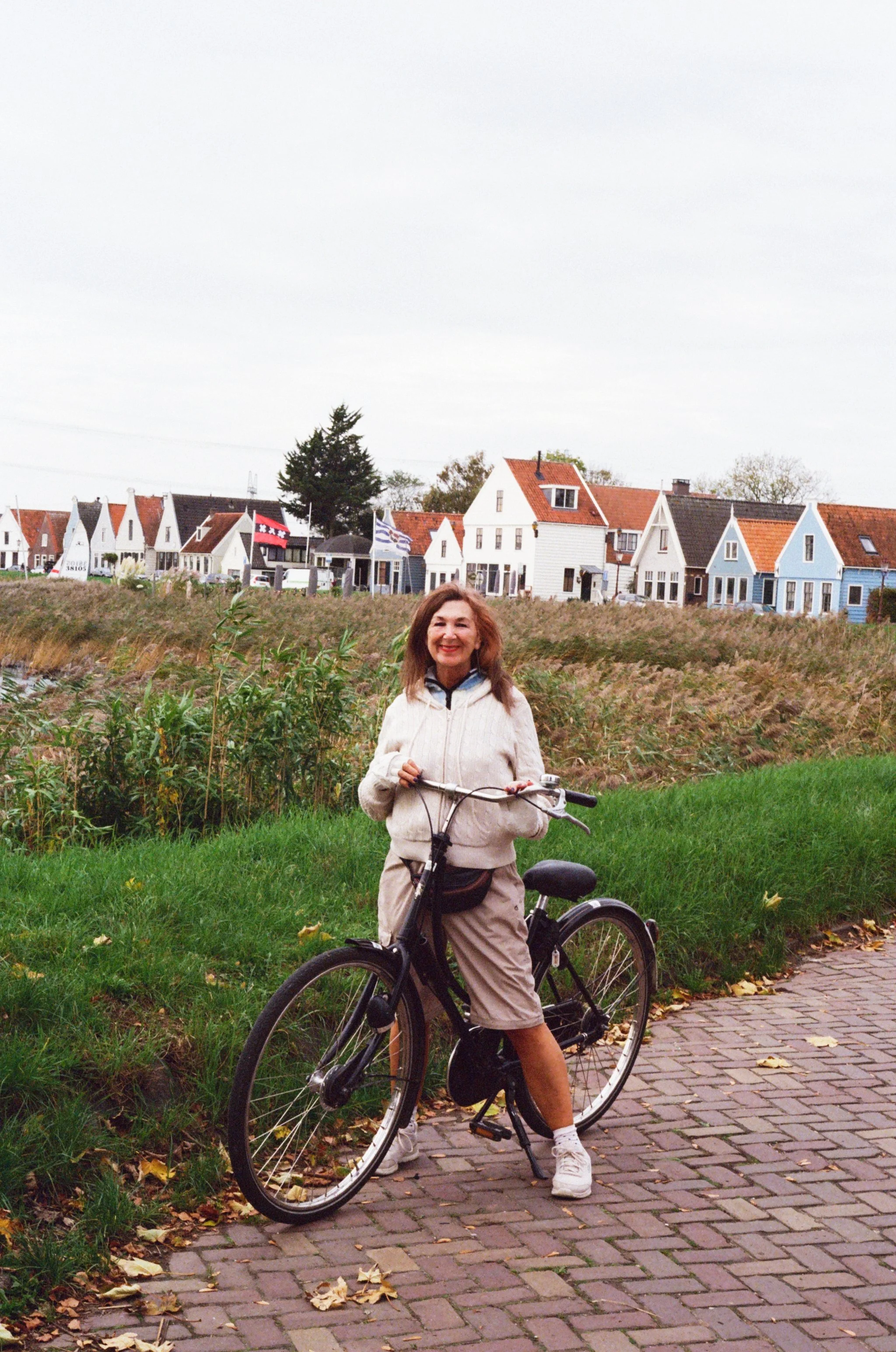 A woman in beige shorts and a cream jacket standing next to a black bicycle on a brick path with colorful houses and a grassy field in the background.