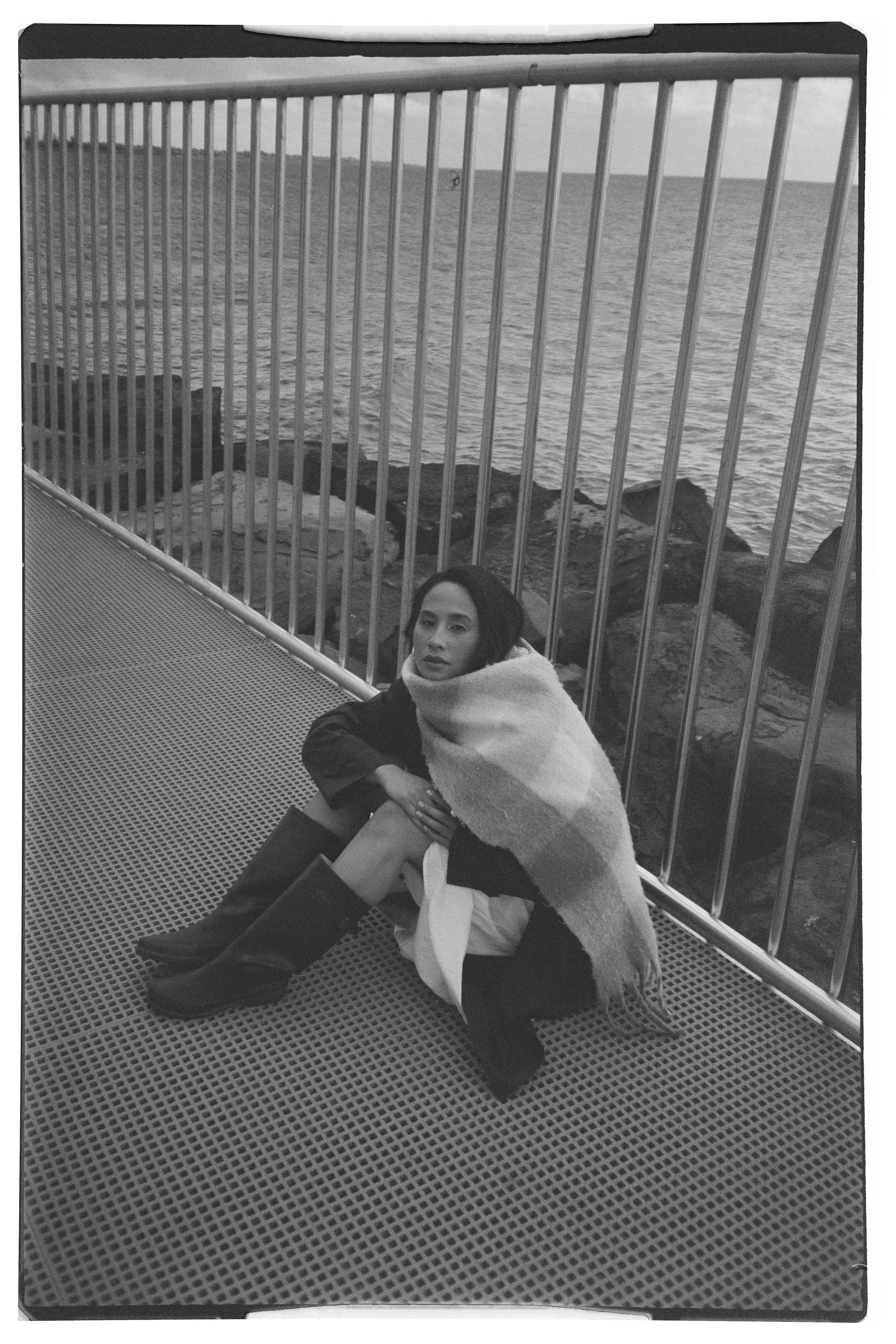 A woman in black boots and a dark jacket sitting on a metal grid surface by the water, wrapped in a large plaid blanket, with a metal railing and rocky shoreline in the background.