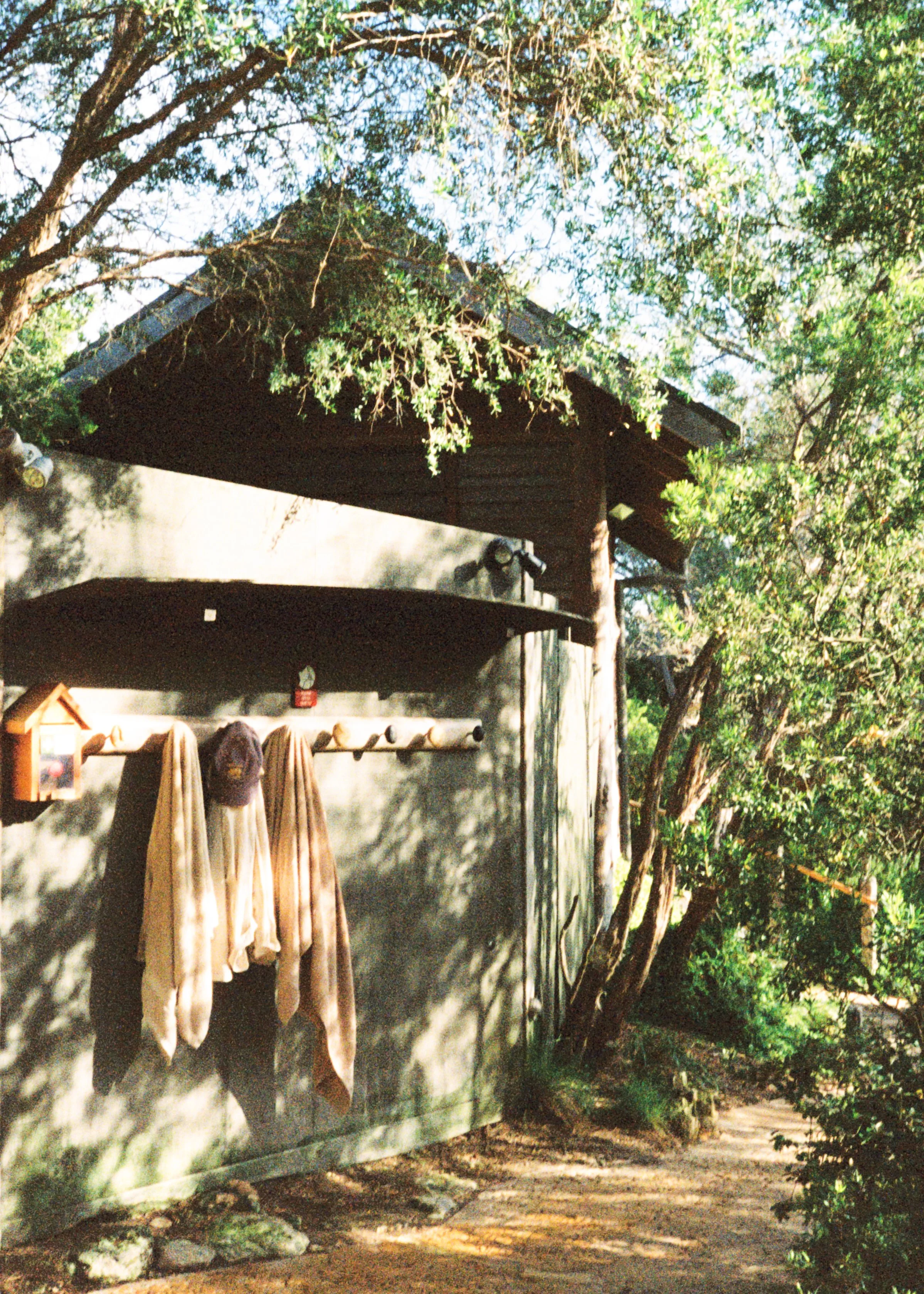 An outdoor scene featuring a small, rustic building with a sloped roof, surrounded by trees and greenery. Two towels or blankets hang on a wooden rack in front of the building, and a pathway runs alongside it.