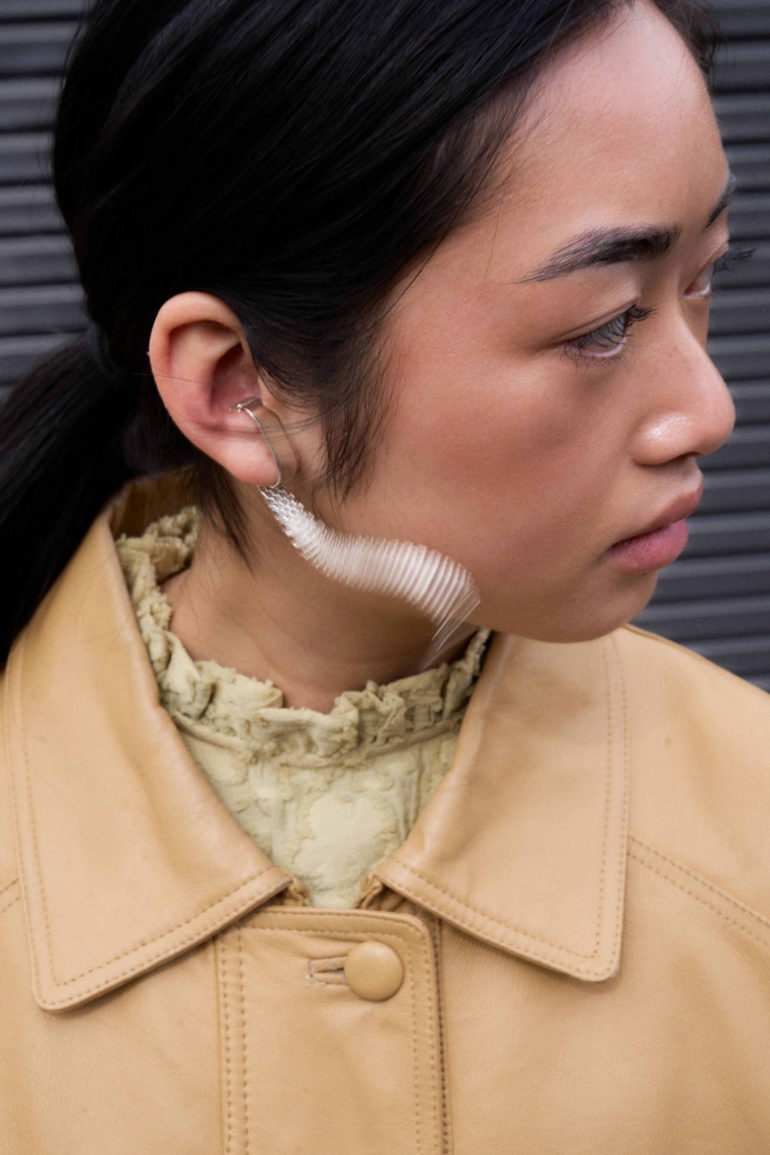Close-up of a woman with dark hair and light brown skin, wearing a beige leather jacket and a ruffled high-neck top, with a transparent, curved plastic earring. The background features dark horizontal lines.