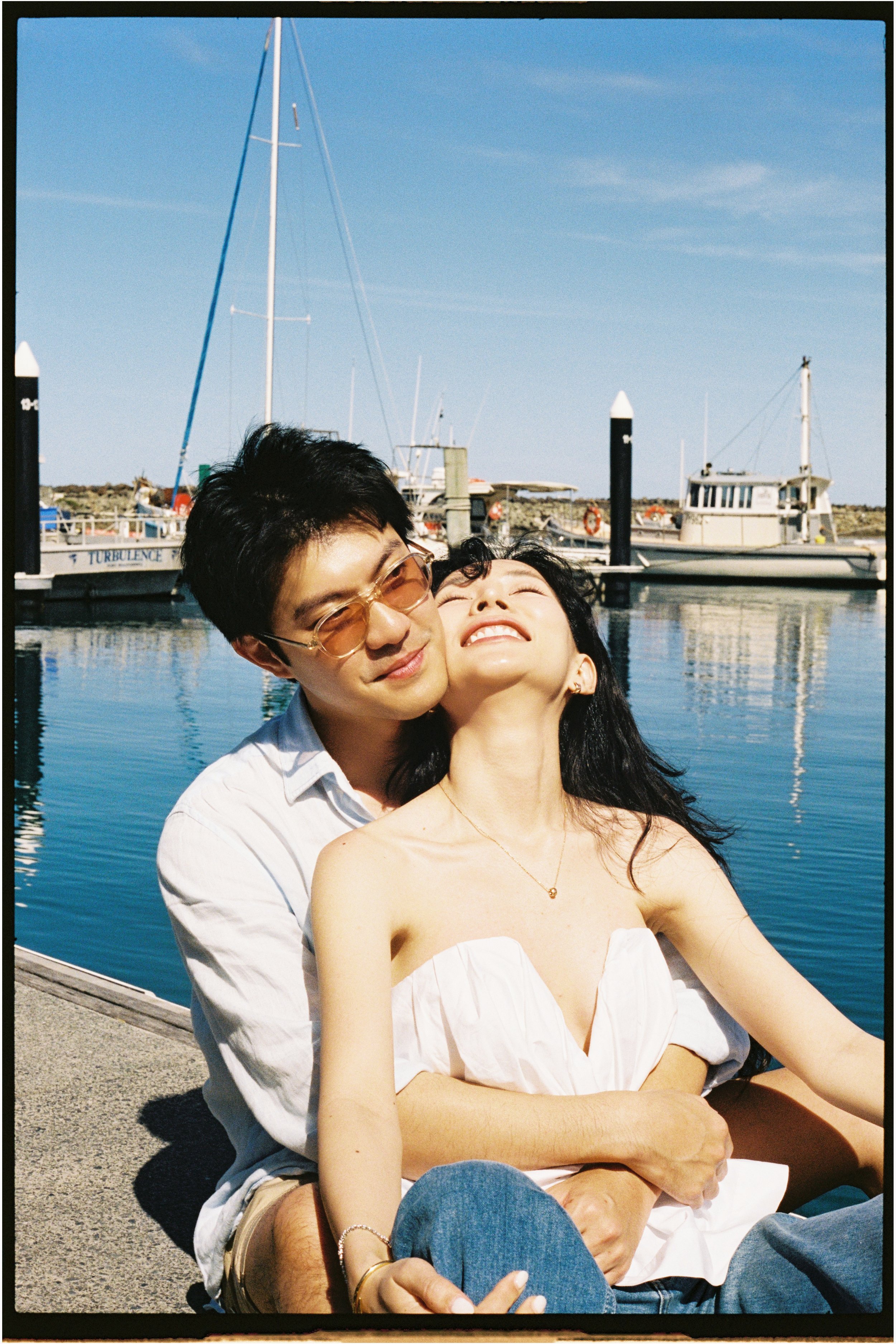 A young couple sitting by a marina on a sunny day, smiling and embracing, with sailboats and calm water in the background.