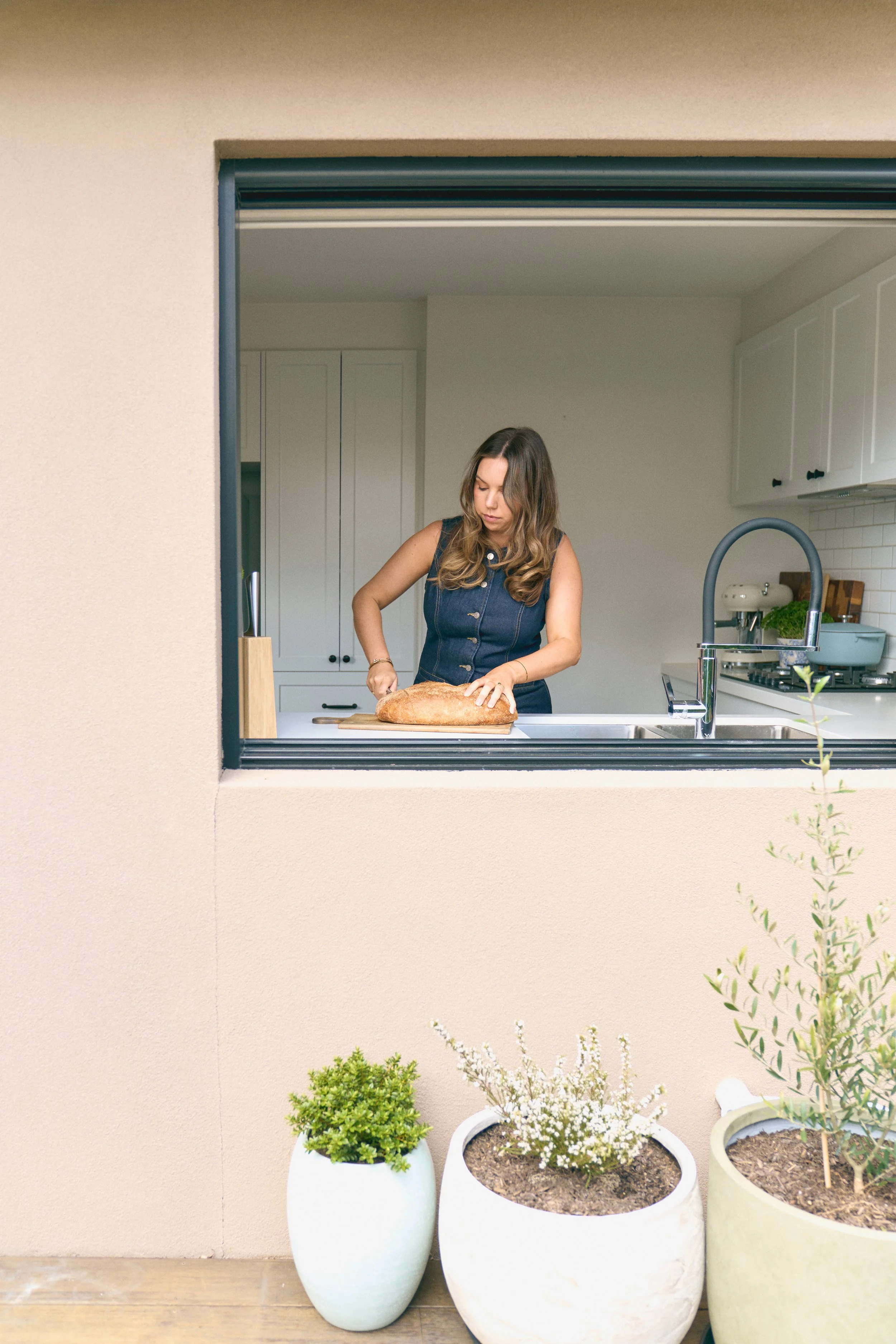 Woman cutting a loaf of bread in a kitchen seen through a window. Outside, potted plants are on a wooden surface.