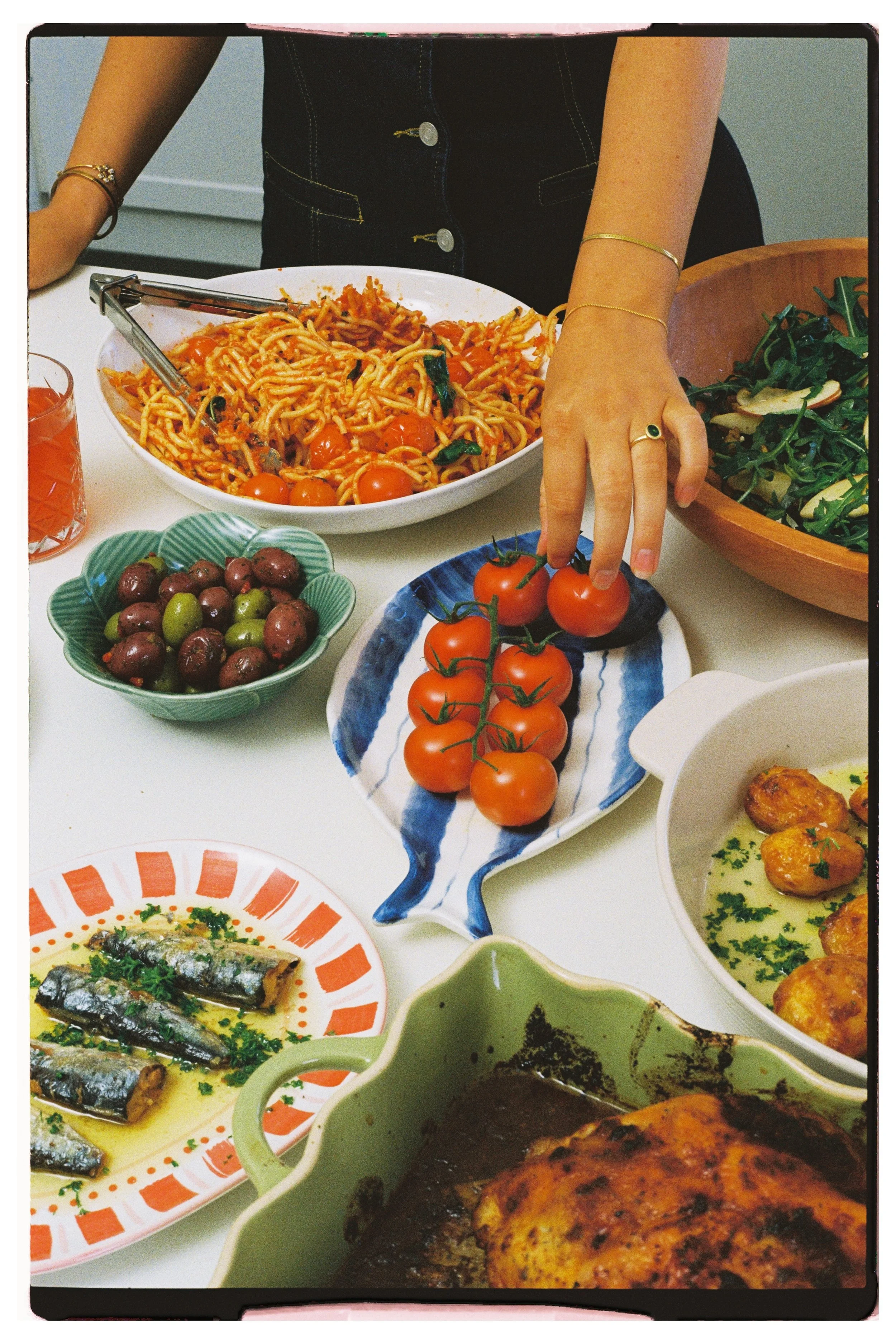 A person reaches for cherry tomatoes on a decorative platter at a meal with various dishes including pasta, fish, and salads.