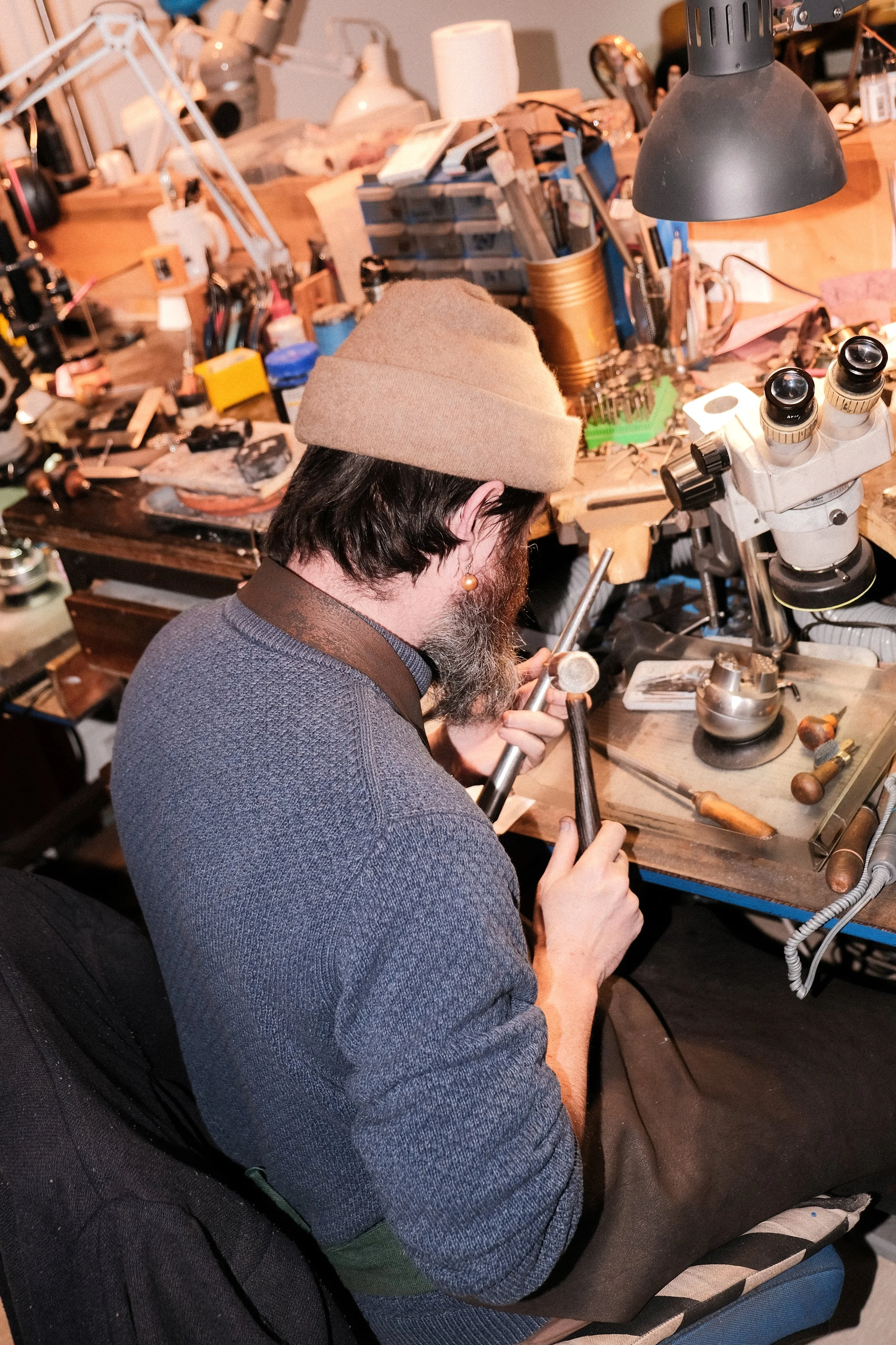 A man in a beige hat and gray sweater working with tools at a cluttered workbench, possibly in a workshop or jewelry-making studio.