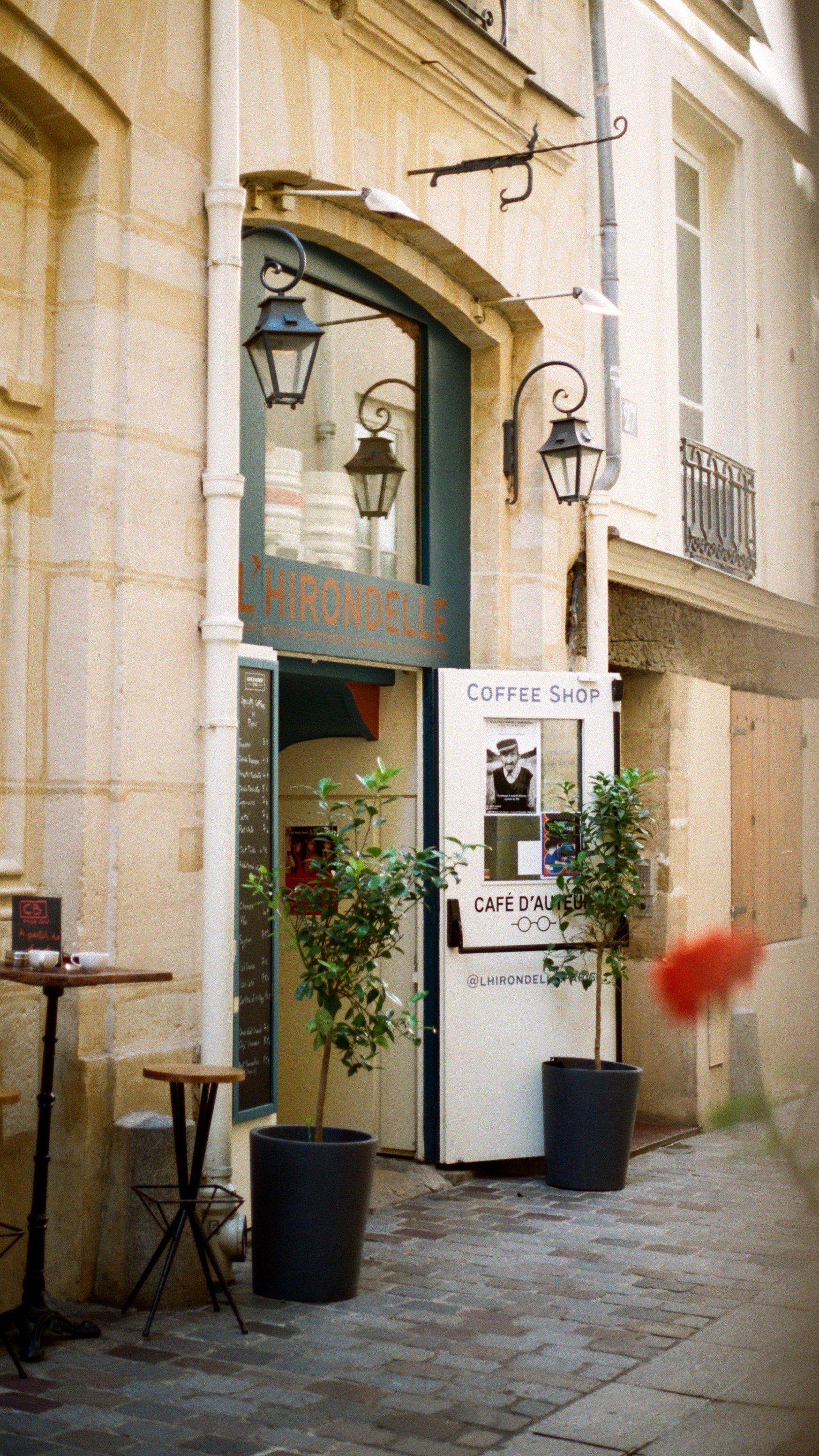 The entrance to a coffee shop called 'LHIRONDELLE' with outdoor seating, potted plants, and decorative lantern-style lights on a stone building.