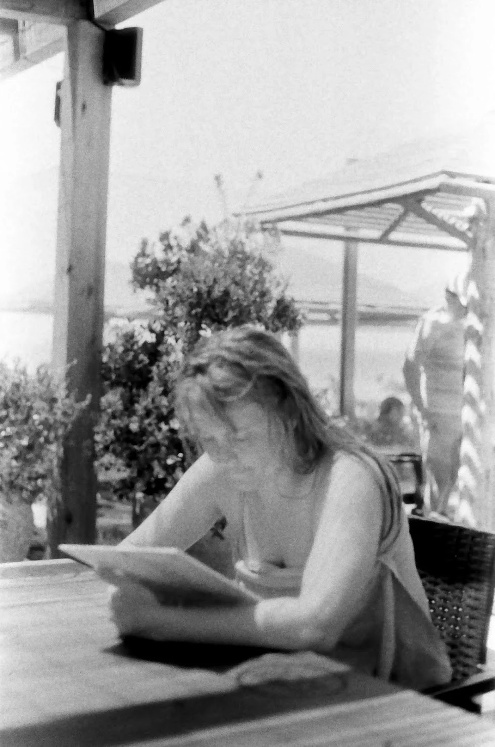 A woman with light-colored hair sitting at an outdoor table, reading a book or tablet. There is a man in the background walking near a structure with a roof, and some potted plants are visible.