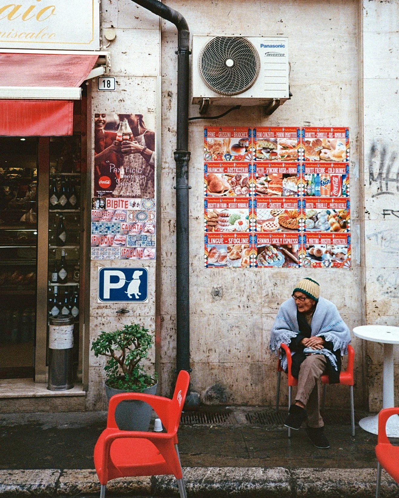 An elderly woman sitting outside a restaurant, with a potted plant, a red chair in the foreground, and a wall displaying a menu with pictures of Italian food. She is wrapped in a blanket and wearing a knit hat.
