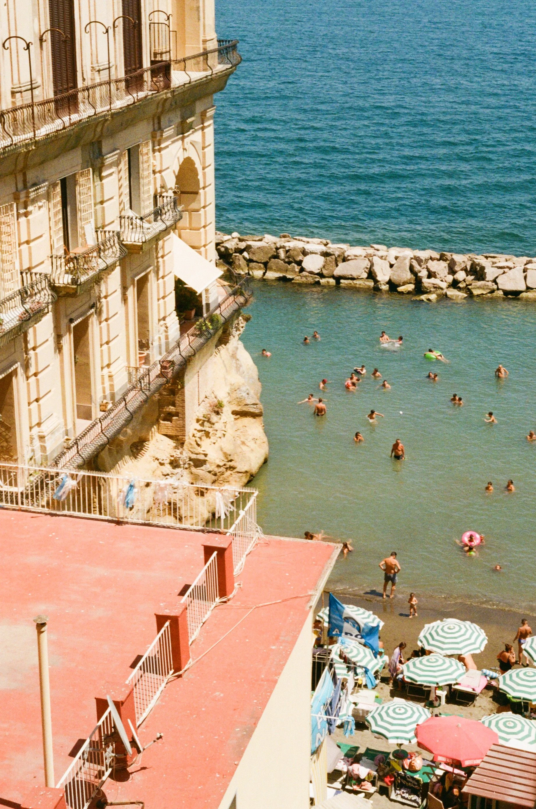 People swimming and relaxing in a small bay next to a building with balconies, with beach umbrellas and lounge chairs nearby.