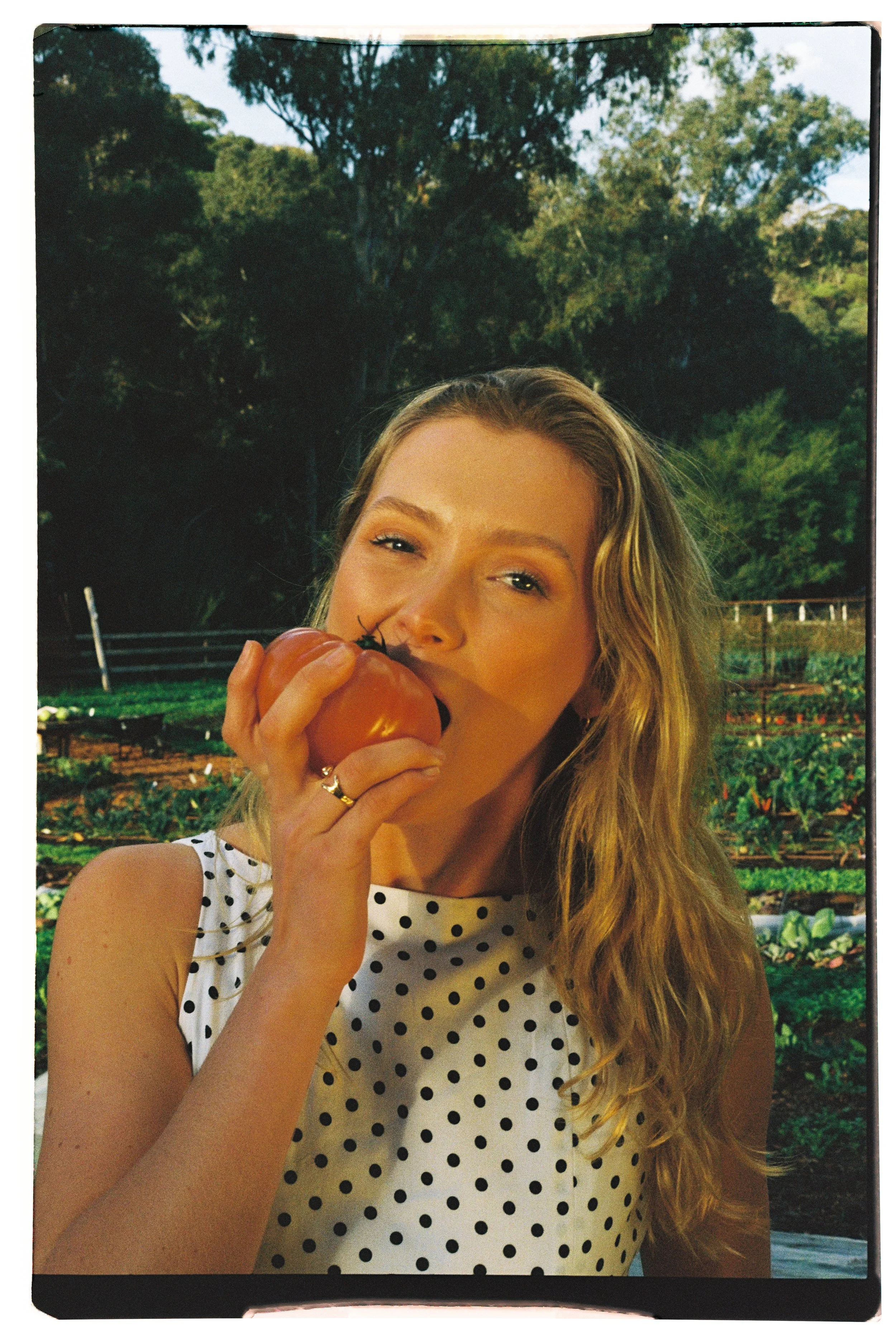 A young woman with blonde wavy hair biting into a large ripe tomato outdoors in a garden with trees and plants in the background.