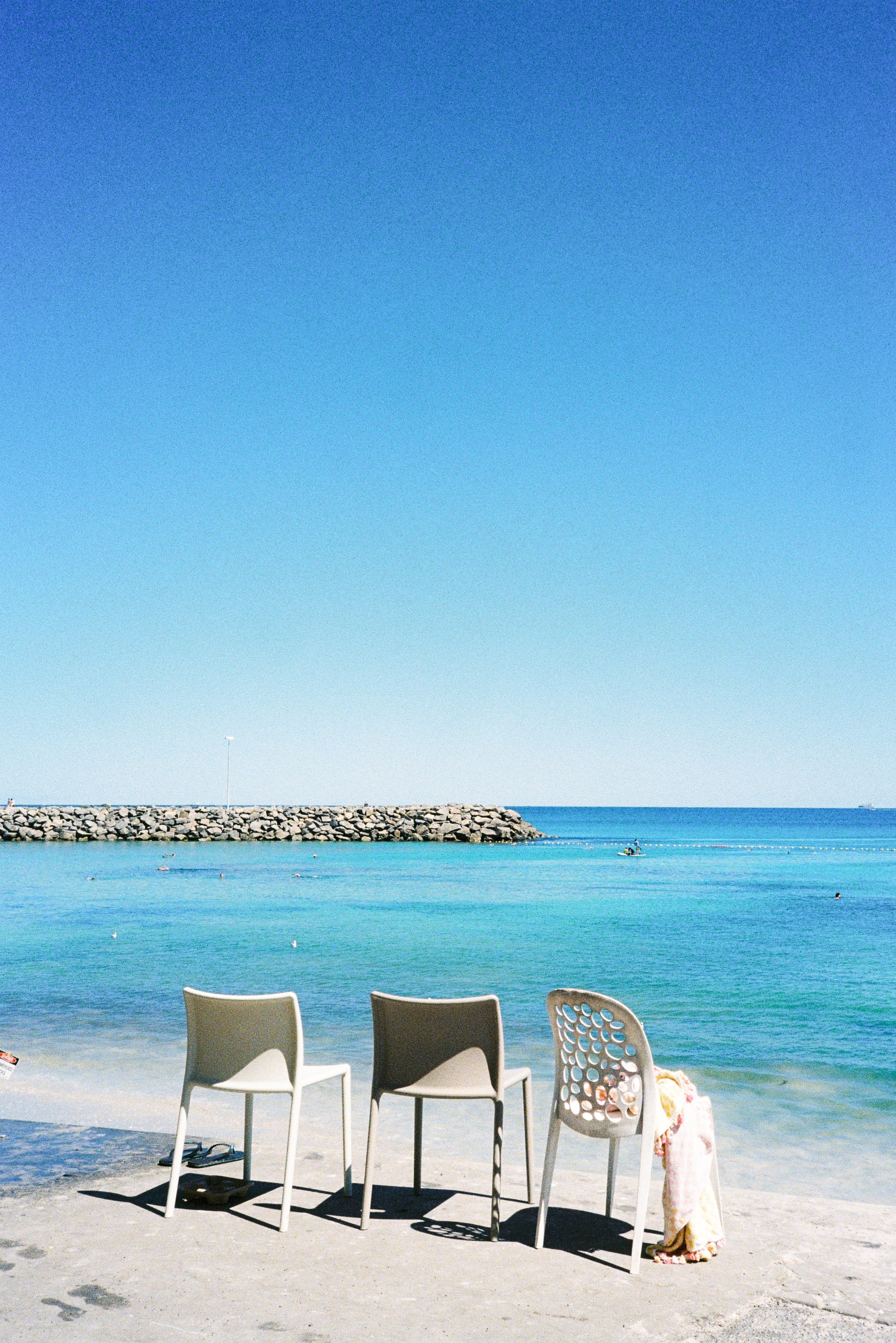 Three chairs on a beach facing the ocean with a breakwater and a boat in the distance under a clear blue sky.