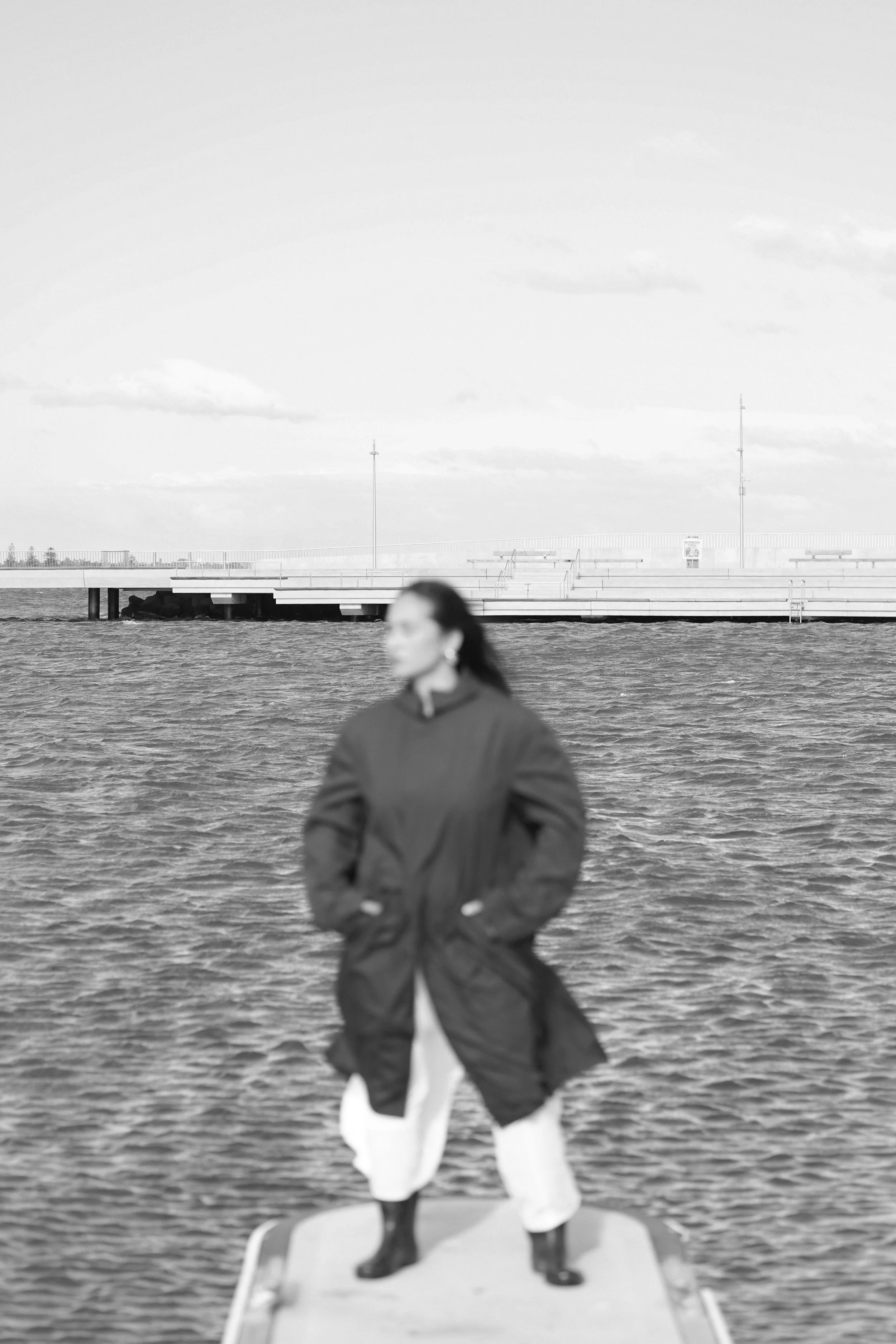 A woman standing on a dock near the water with her hands in her coat pockets, wearing rain boots and a coat, with a city skyline and an empty pier in the background.